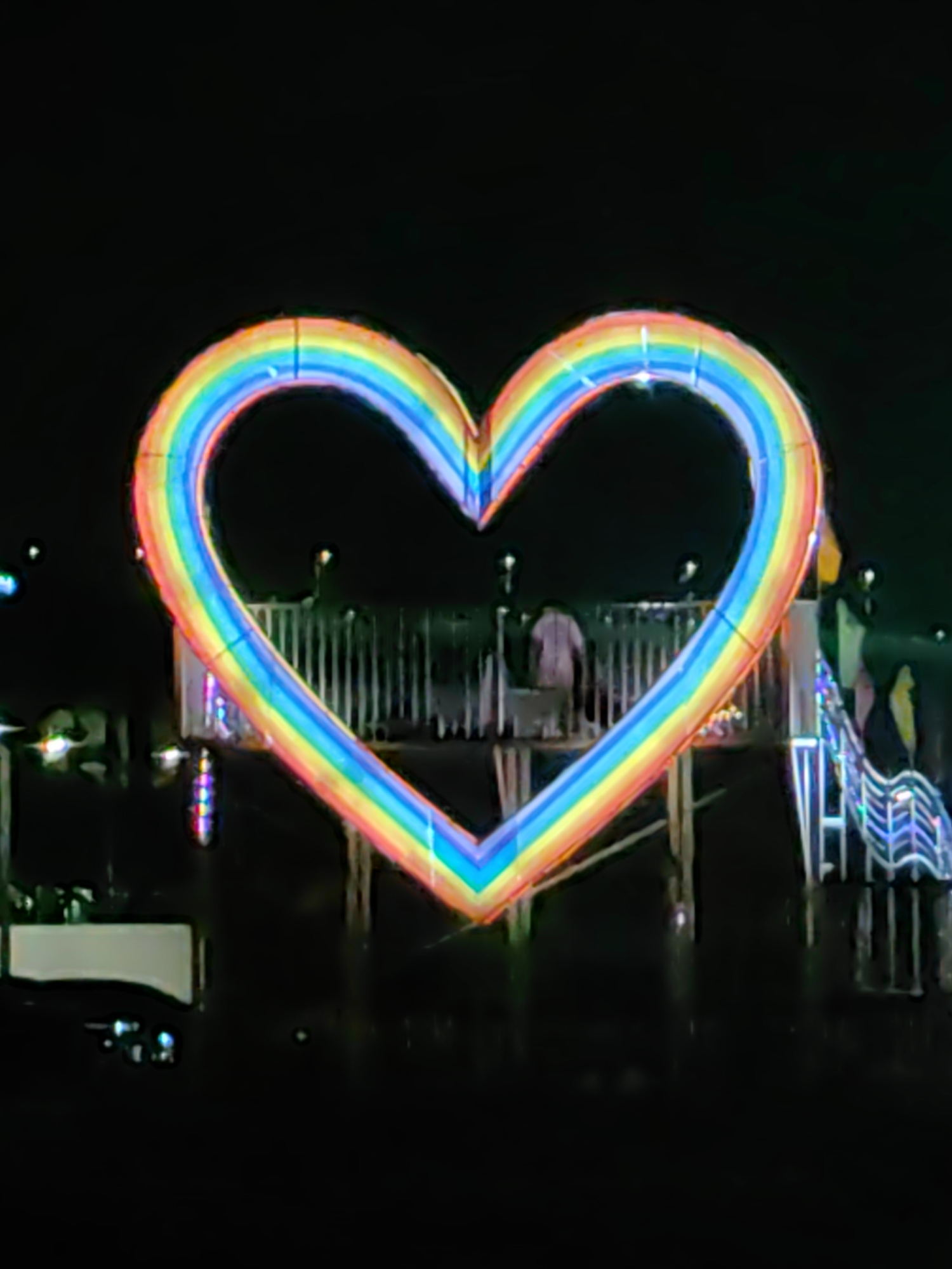 At night in Bai Chay Ward, Vietnam, a large, illuminated rainbow-colored heart sculpture stands on a structure over water. Faintly visible behind the heart are people and railings. The water reflects some of the lights. Other illuminated structures are in the background, though the scene is primarily dark.