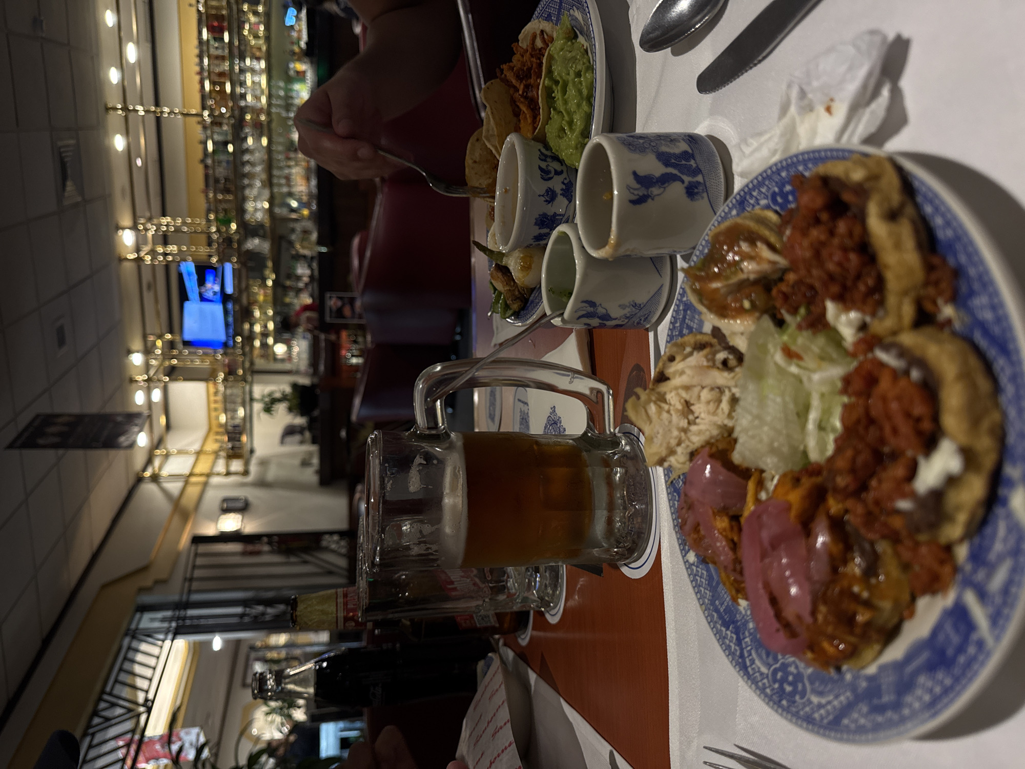 The image, rotated ninety degrees clockwise, captures an indoor dining scene at a restaurant or bar, likely in the evening. In the foreground, a wooden table with a white placemat is laden with a variety of Mexican dishes and drinks.
The main subject is a blue and white patterned plate filled with antojitos, possibly sopes or similar corn-based snacks. These are generously topped with various ingredients: what appears to be shredded chicken, a red meat mixture (possibly chorizo or cochinita pibil), fresh lettuce, white cream or cheese, and vibrant pickled red onions. Next to this, another plate holds tacos with a meat filling and a portion of bright green guacamole. Small, matching blue and white ceramic dishes contain accompanying sauces or grilled vegetables, including what looks like grilled onions and peppers.
A large glass mug filled with a dark amber beverage, likely beer, showing some foam, is prominently placed. A dark glass bottle, possibly Coca-Cola, is partially visible nearby. Silverware, including forks, spoons, and a knife, rests on the white placemat. A person's hand holding a fork reaches towards the food on the left.
The background reveals the lively interior of the establishment. A brightly lit bar area with extensive shelving displaying numerous bottles of liquor stretches across the midground. The lighting is warm and artificial, suggesting a nighttime setting. A television screen, glowing blue, is visible on a distant wall. The overall ambiance appears casual and inviting, consistent with a dining experience in Puebla, Mexico, given the traditional Mexican cuisine on display. No legible text is clearly visible on any object.