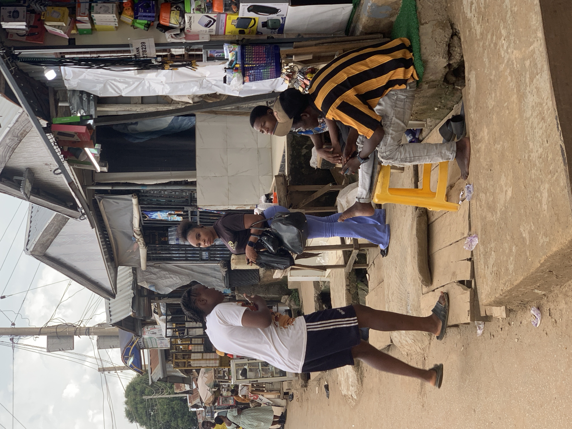 In Abuja, Nigeria, this image captures a street scene near an outdoor market. A young man with braided hair, wearing a white t-shirt and dark shorts, stands and gestures as he talks to a woman in a dark t-shirt and light blue flared pants. The woman holds two black bags. To their right, two young men are seated on a yellow plastic stool and a raised concrete platform. One man, wearing a black and orange striped t-shirt and light grey ripped jeans, is looking away, holding a mobile phone. The other man, wearing a t-shirt and a cap, also looks away. The background shows stalls with various goods displayed, including electronics, and a cloudy sky. The ground is dusty and uneven, with some wooden planks and concrete structures.
