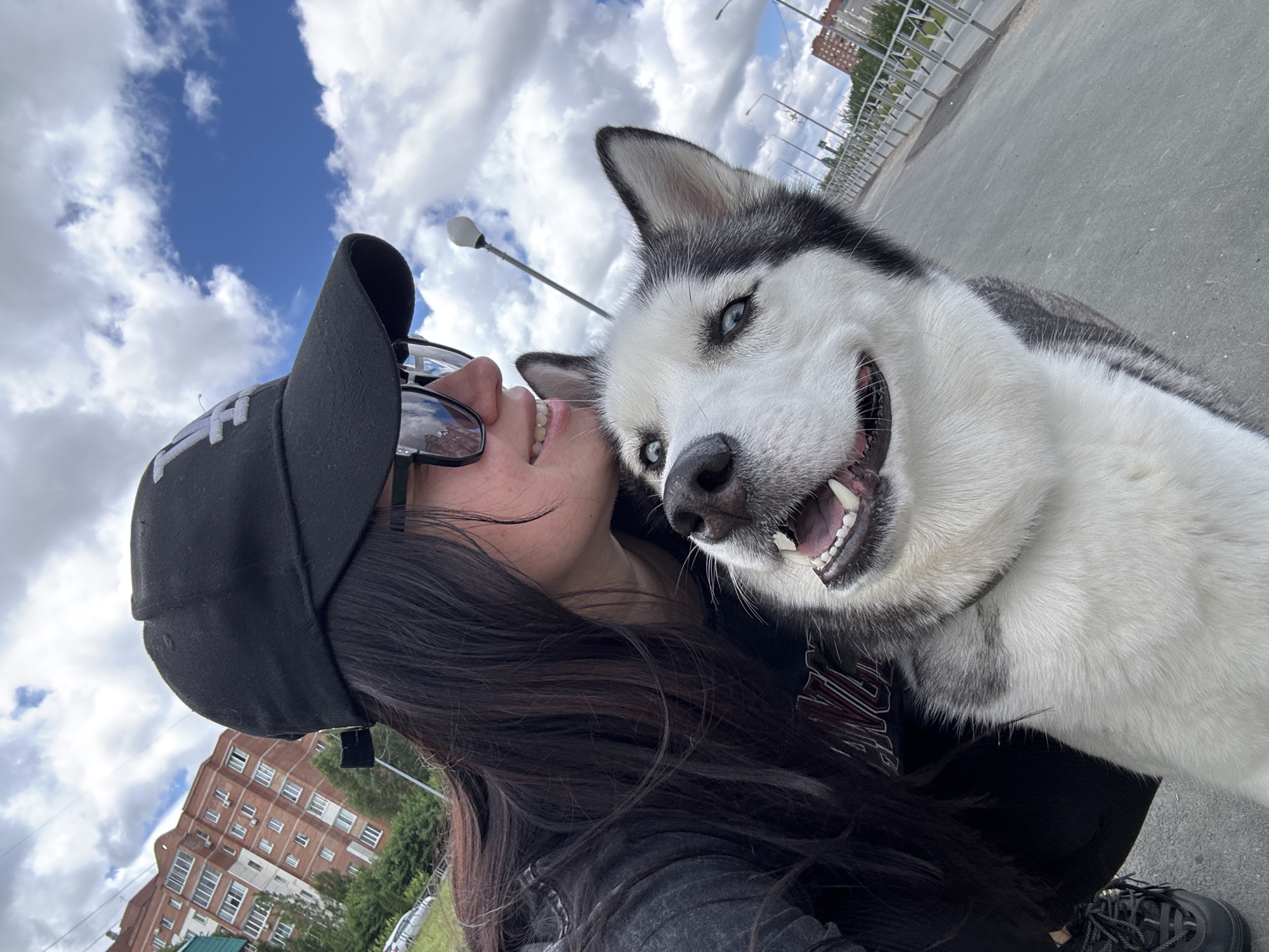 A joyful outdoor selfie captures a person and their Siberian Husky. The image is taken at a slight angle, with the subjects appearing from the lower left, looking up towards the camera.
The person, visible from the chest up, has long dark hair and is wearing a black baseball cap with white embroidered text, possibly "TH" or "H", on the front. They also wear dark sunglasses perched on their head and a dark top or jacket with an indistinct design. A bright smile is visible on their face as they look towards their dog.
Beside the person, a striking black and white Siberian Husky dominates the right side of the frame. The dog has piercing blue eyes and a wide, open-mouthed grin, revealing its teeth and tongue, conveying a highly enthusiastic and happy expression as it looks directly at the camera. Its fur is a mix of white and dark grey/black, typical of the breed.
The setting is an outdoor urban or suburban environment. The background features a partly cloudy blue sky, with white clouds scattered across. In the distance, there are multi-story brick buildings, possibly apartment blocks, and light poles with lamps. A concrete road or pathway stretches behind the dog, with a metal barrier or railing visible further back. The overall mood is cheerful and affectionate, capturing a happy moment between the person and their pet on a bright day.