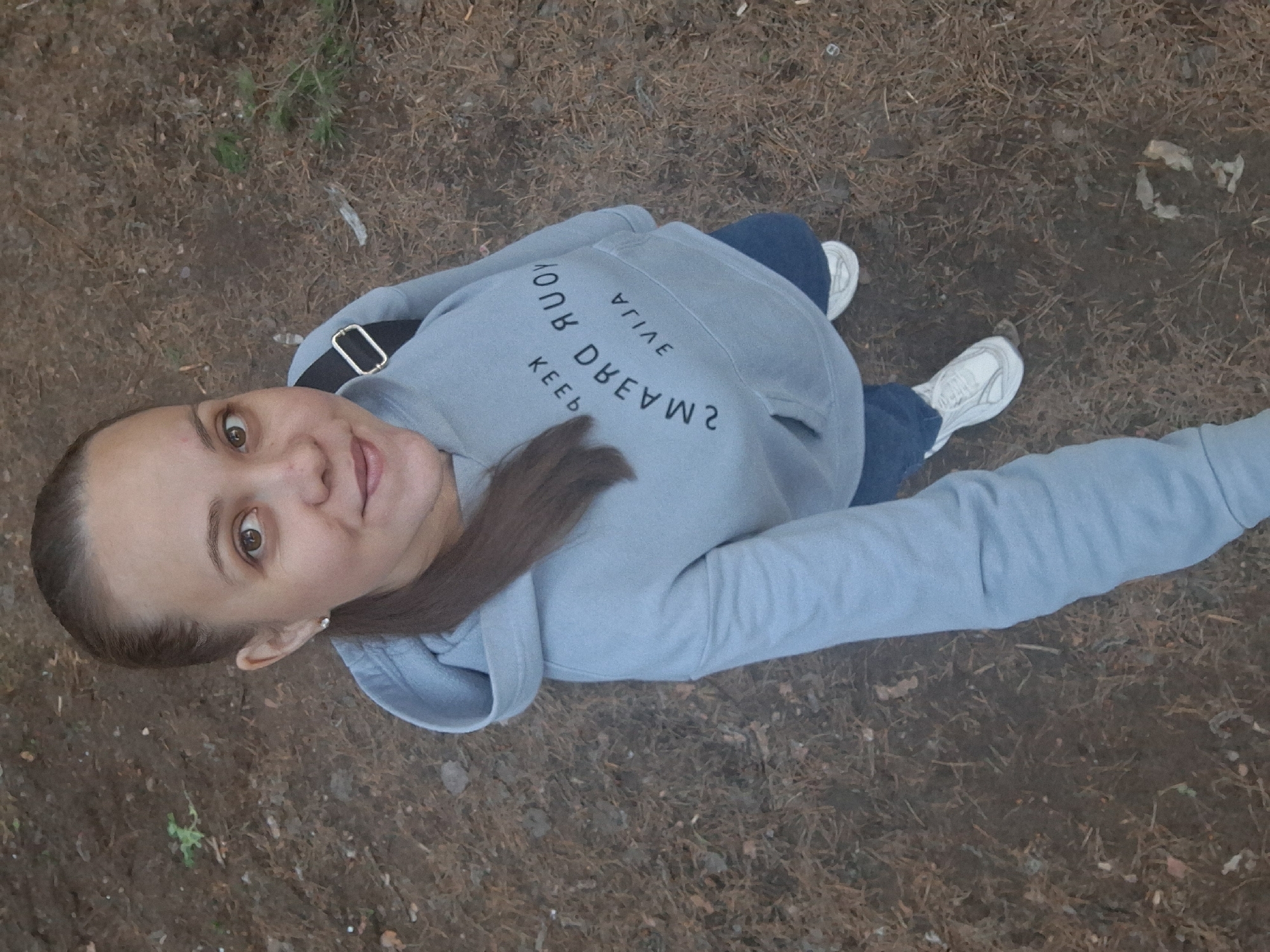 A woman stands in an outdoor setting, seemingly a forest or wooded area, as evidenced by the pine needles and leaves covering the ground. She is wearing a light blue hoodie with text on the back, blue jeans, and white sneakers. She is looking up at the camera, smiling slightly. The photograph appears to be taken from an elevated angle, looking down at the subject. She is also wearing a strap of some kind across her body. The location is Chaikovsky, Russia.