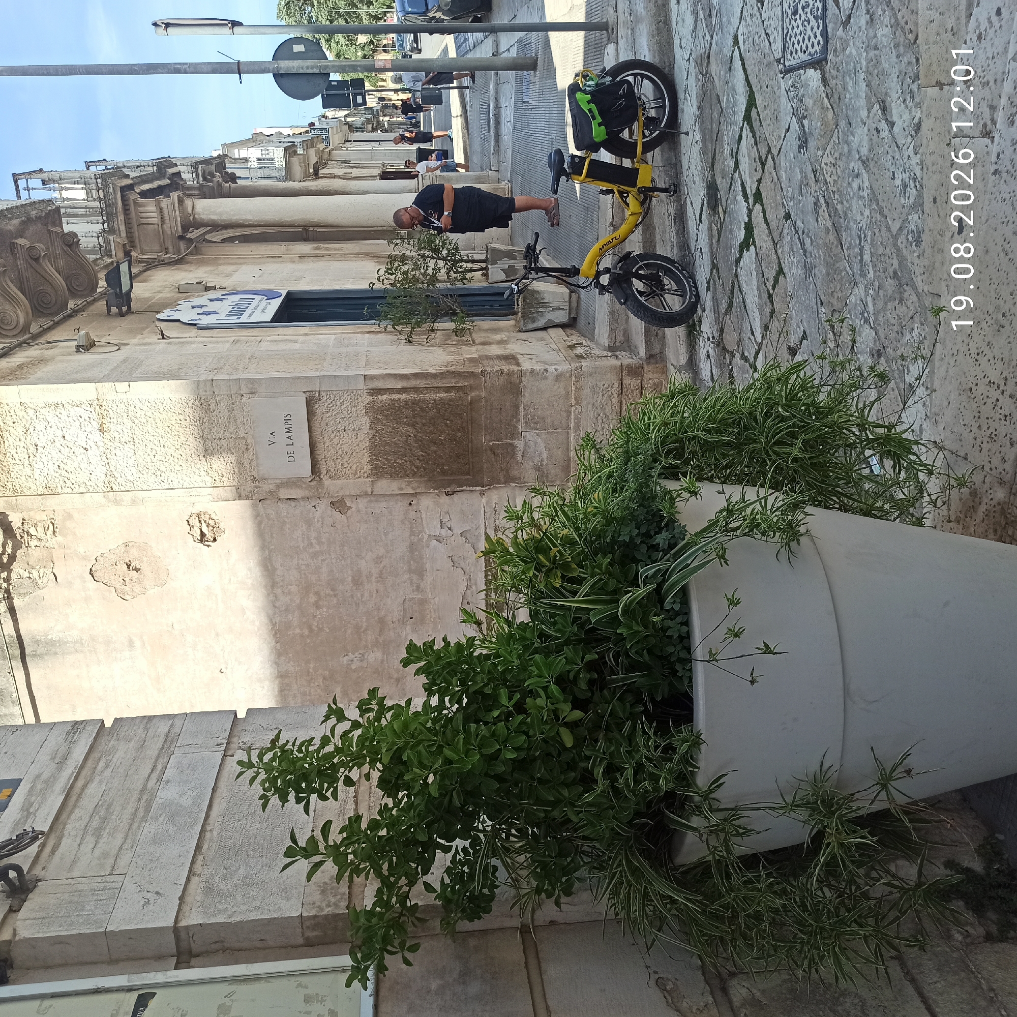 The image depicts a street scene in Ruvo di Puglia, Italy. In the foreground, a large white planter contains dense green foliage. To the right, a yellow electric bicycle is parked on a cobblestone sidewalk. A man wearing a black t-shirt and shorts stands near the bicycle, looking at his phone. In the background, a building with ornate architecture lines the street. A sign on the building reads "VIA DE LAMPIS". The sky is blue and clear. The overall impression is of a sunny day in a historic Italian town.