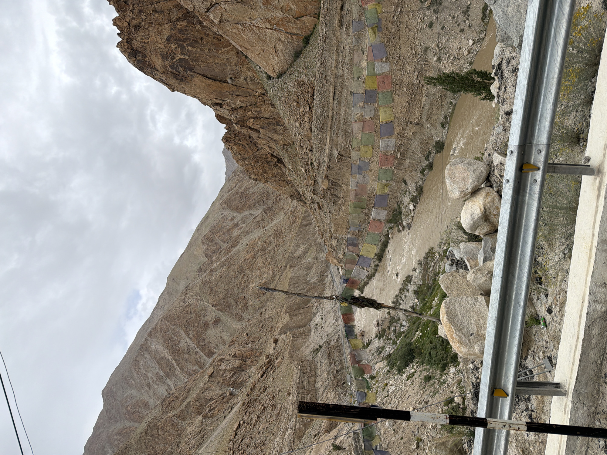 An eye-level, long shot captures a wide, brown river flowing through a narrow gorge between steep, barren, brown-and-tan rocky mountains under a cloudy sky. Prayer flags in various colors, including red, yellow, blue, green, and white, are strung along the side of the gorge, leading down to the river. A single, thin, dead tree trunk with some fabric tied to it stands vertically in the foreground. On the left, a black and white striped pole is visible. The foreground features a grey metal guardrail above a concrete barrier, with large rocks and some scrubby vegetation. The image appears to be taken from a roadside vantage point. The location is identified as Gayk, India.