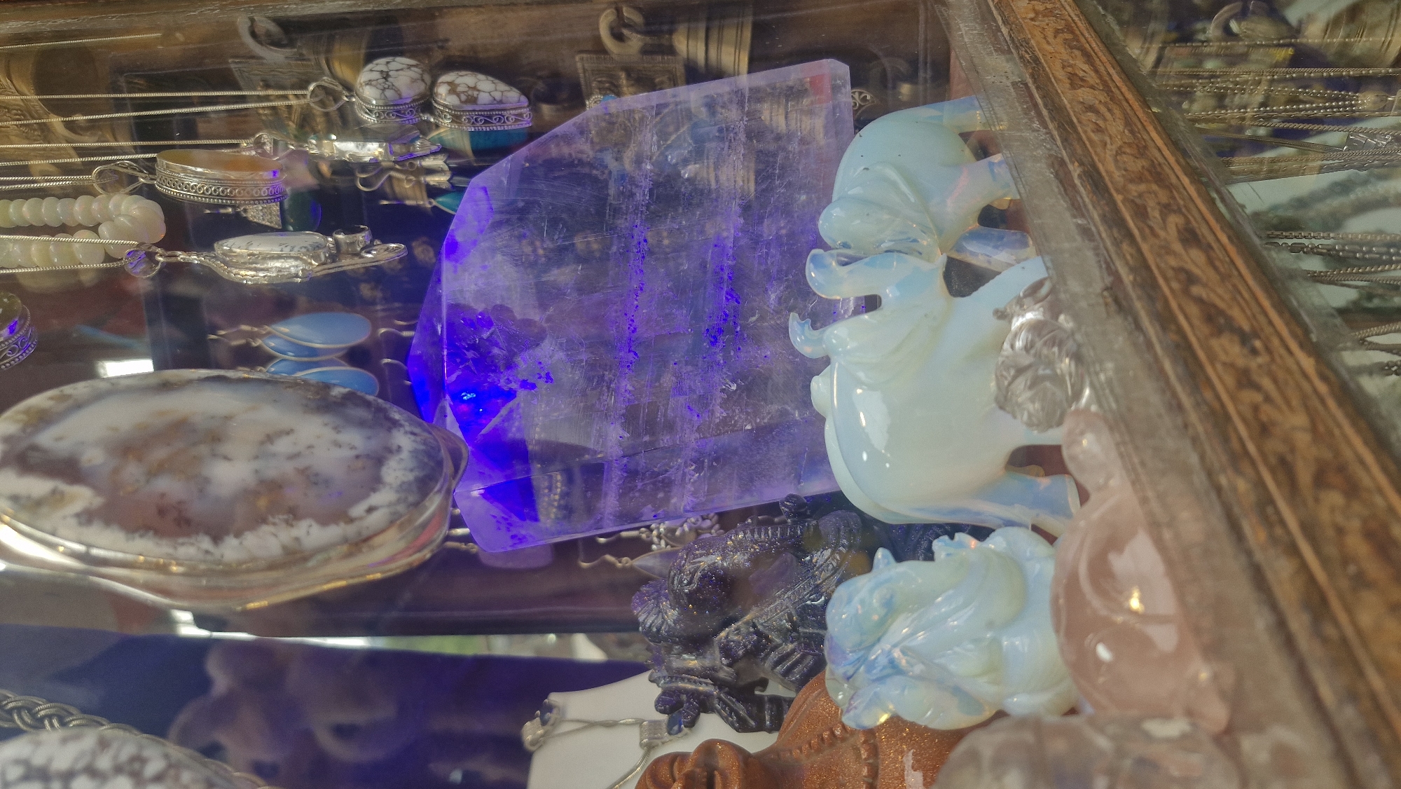 This is a close-up, eye-level, interior shot of a display case in Vashisht, India, filled with various decorative and spiritual items. The lighting is somewhat dim with a strong purple hue emanating from behind a large, translucent crystal slab. In the foreground, on a wooden shelf, are two pale blue, translucent elephant figurines. To the left of the elephants is a carved dark purple figurine, possibly Ganesha. Behind the elephants and to the left, a portion of a reddish-brown figurine with a smiling face is visible.
Above these figurines, suspended from above, are numerous pieces of jewelry. These include silver chains, pendants with stones, and earrings. A large, oval-shaped pendant with a marbled brown and white pattern is prominently displayed in the upper left quadrant. To the right of this, several teardrop-shaped blue earrings are visible.
The background is cluttered with more hanging jewelry and various objects, including what appears to be a large, uncut crystal or geode in the center, illuminated by the purple light. The overall impression is a retail display, likely in a shop or market stall.