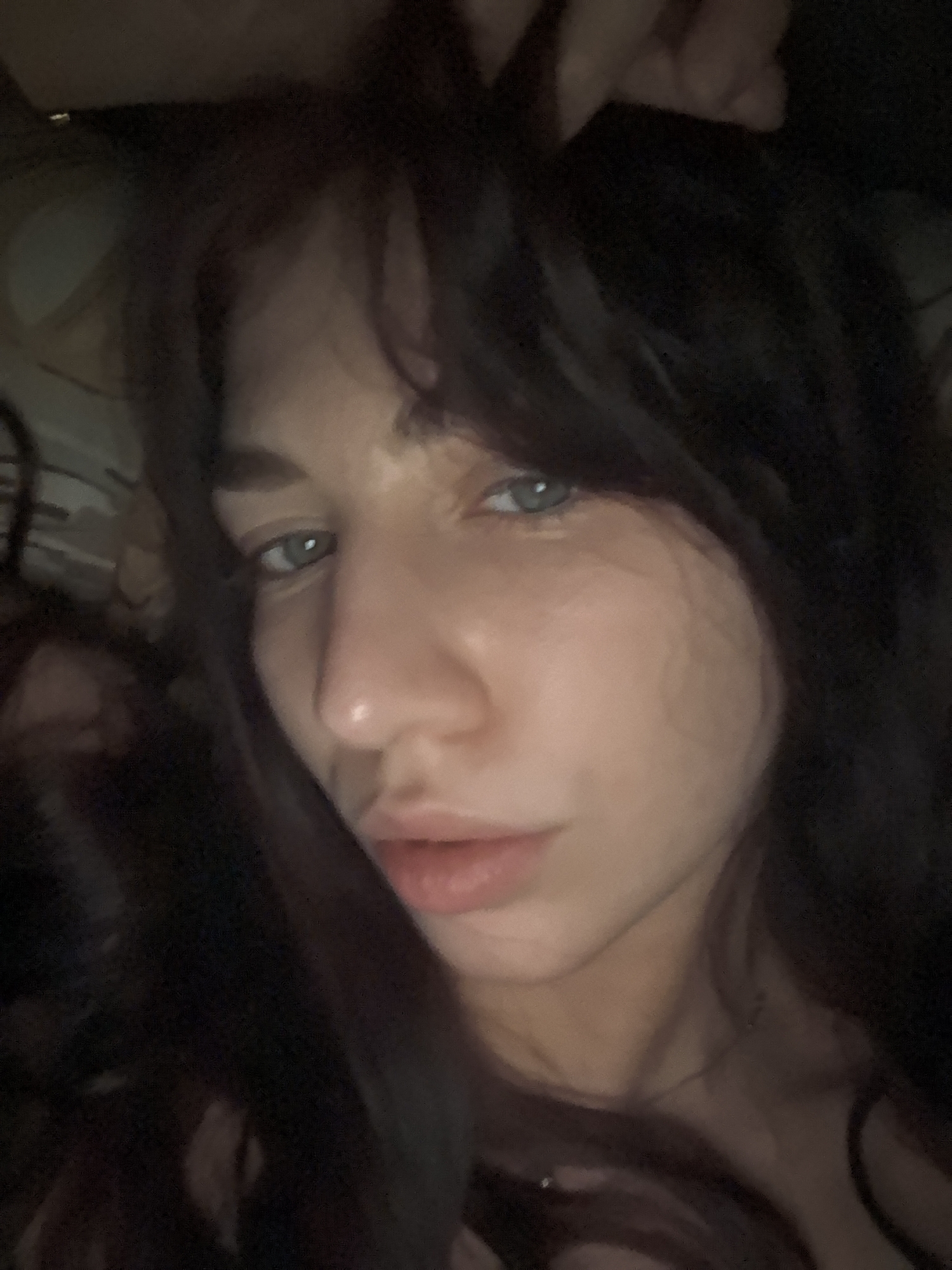 A close-up, dimly lit photograph shows a person with light skin, dark brown hair with bangs, and light-colored eyes. The person's face is the primary focus, captured at a slight angle. The lighting is uneven, casting shadows across the face and hair. There are no visible details to indicate the location or city.