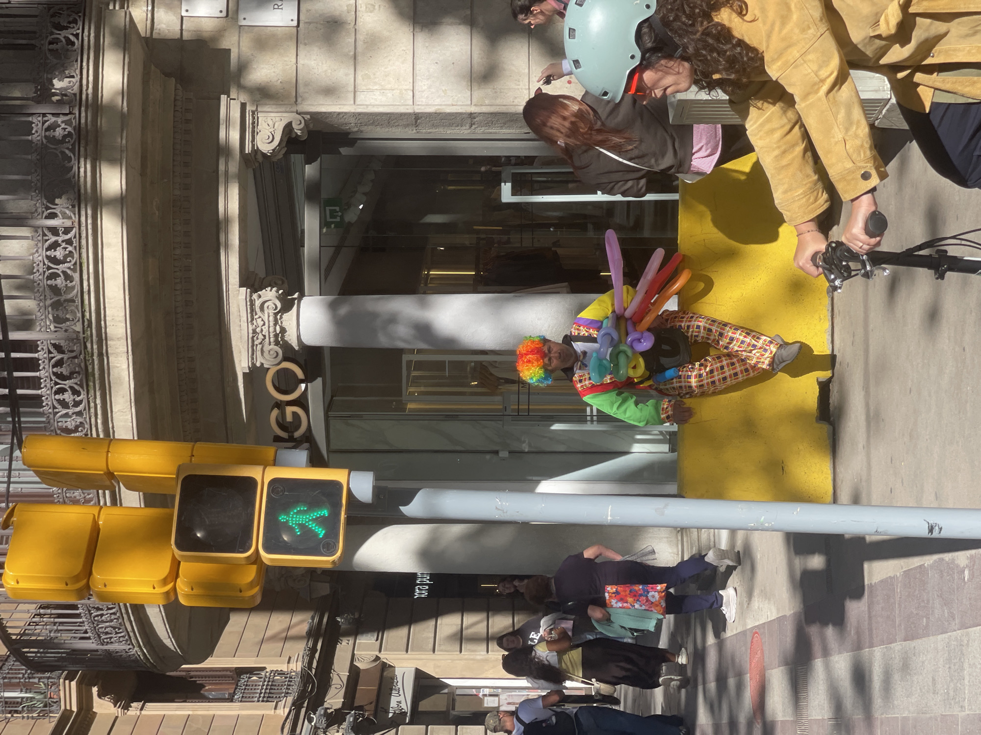 The image captures a street scene in Barcelona, Spain. A clown wearing a colorful wig, yellow and green jacket, and checkered pants is seated on a yellow curb, surrounded by an assortment of balloons. Adjacent to the clown is a woman on a bicycle, who seems to be interacting with the clown. Pedestrians are walking on the sidewalk. There is a traffic light displaying a green walking person. The building in the background has classic architectural details, and the letters "GO" are visible. The overall scene is bright and sunny.