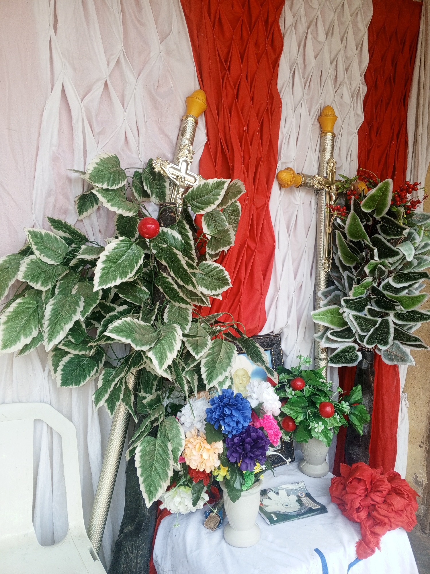 The image captures a makeshift altar in Lokoja, Nigeria. The backdrop consists of draped white and red fabric. Two large, silver-colored crosses with gold-colored finials stand on either side, partially obscured by artificial greenery with variegated leaves and decorative red berries. A white table is covered with a cloth, upon which sits a framed photograph of a man, several vases of colorful artificial flowers, a small white vase containing greenery and red berries, a magazine, and a crumpled red cloth. A white plastic chair is partially visible on the left side of the image. The setting suggests a humble, personal memorial or religious display.