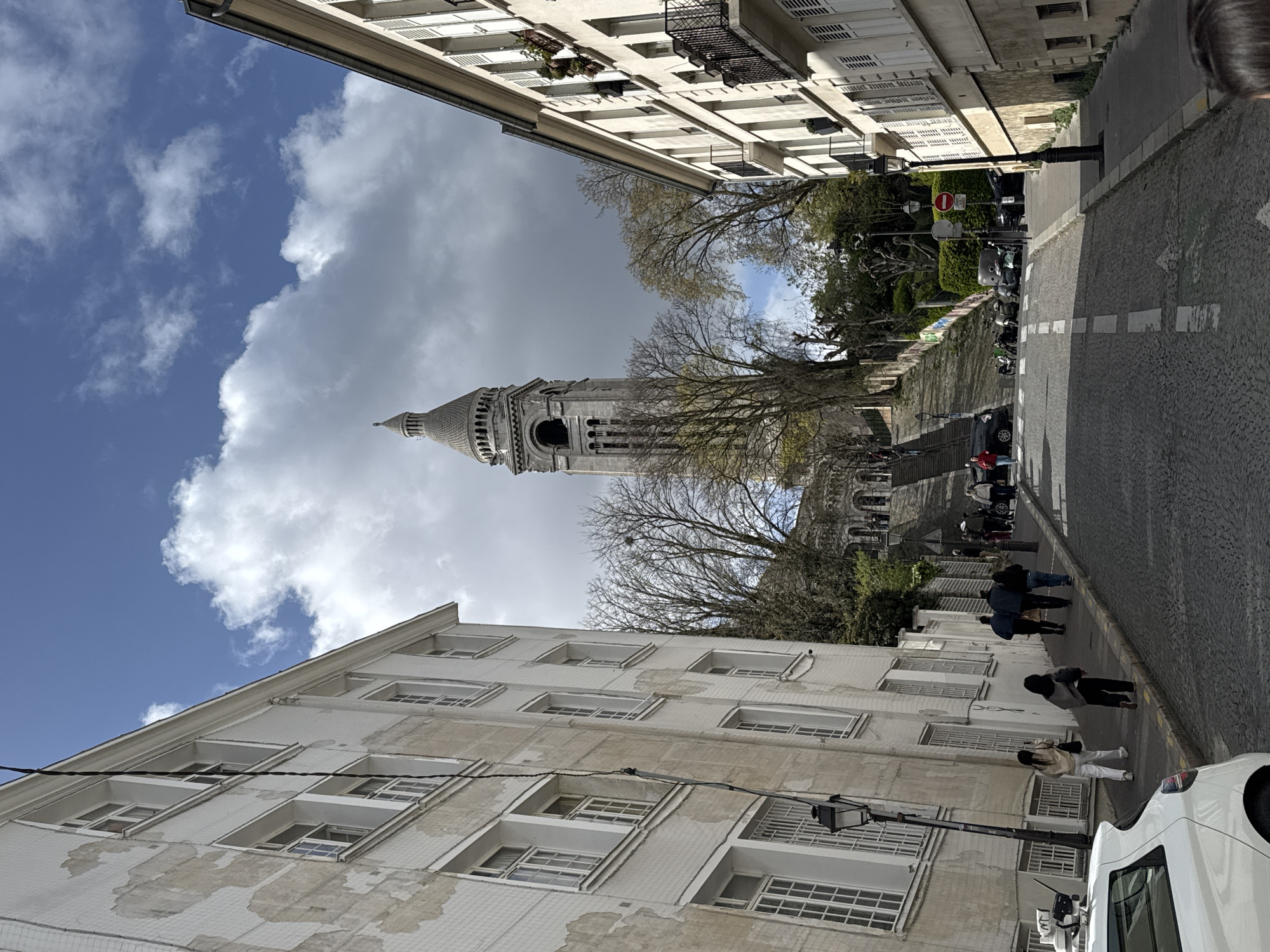 The image captures a street scene in Paris, France, featuring the Sacré-Cœur Basilica visible in the distance. The shot is taken from a low angle, looking up the street towards the basilica. The street is lined with buildings on either side, characterized by multiple windows and classic Parisian architecture. Pedestrians are walking along the street, some on the sidewalk and others on the cobblestone pavement. A white van is parked along the side of the street, near the bottom right of the image. The sky is partly cloudy, with patches of blue sky visible among the clouds. Trees are scattered along the street, adding greenery to the urban landscape. The Sacré-Cœur Basilica stands prominently at the end of the street, its white stone contrasting with the sky. A flight of stairs leading up to the basilica is also visible.