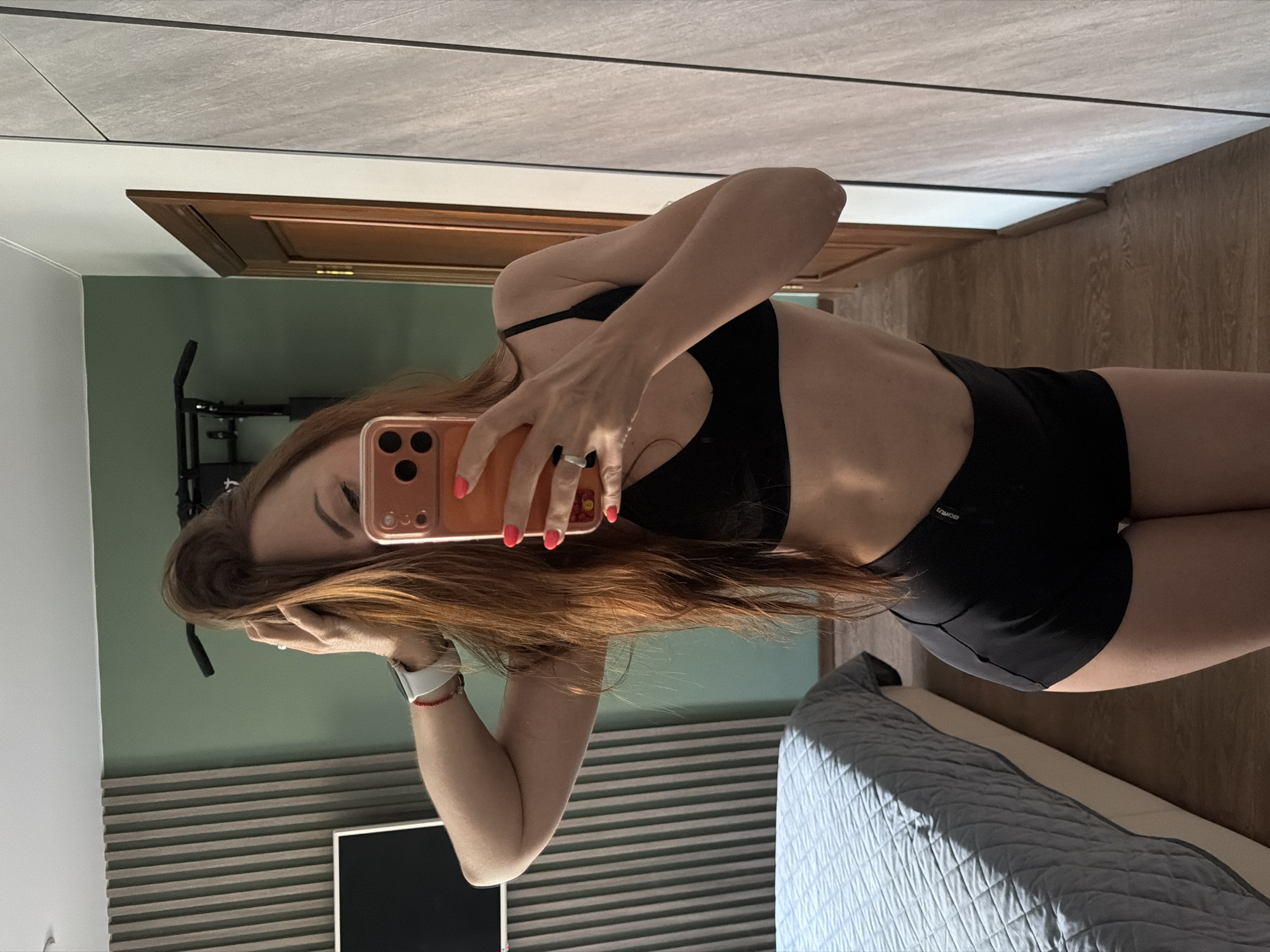 A woman with long, auburn hair is taking a mirror selfie in a bedroom. She is wearing a black sports bra and black shorts. She is holding her phone in her right hand, and her left arm is raised, with her hand in her hair. A bed with a grey quilted comforter is visible to the left of the frame. The room has green walls, and a wooden door and closet are visible to the right. A pull-up bar is mounted on the wall in the background. There is no indication of the location being Birobidzhan, Russia.
