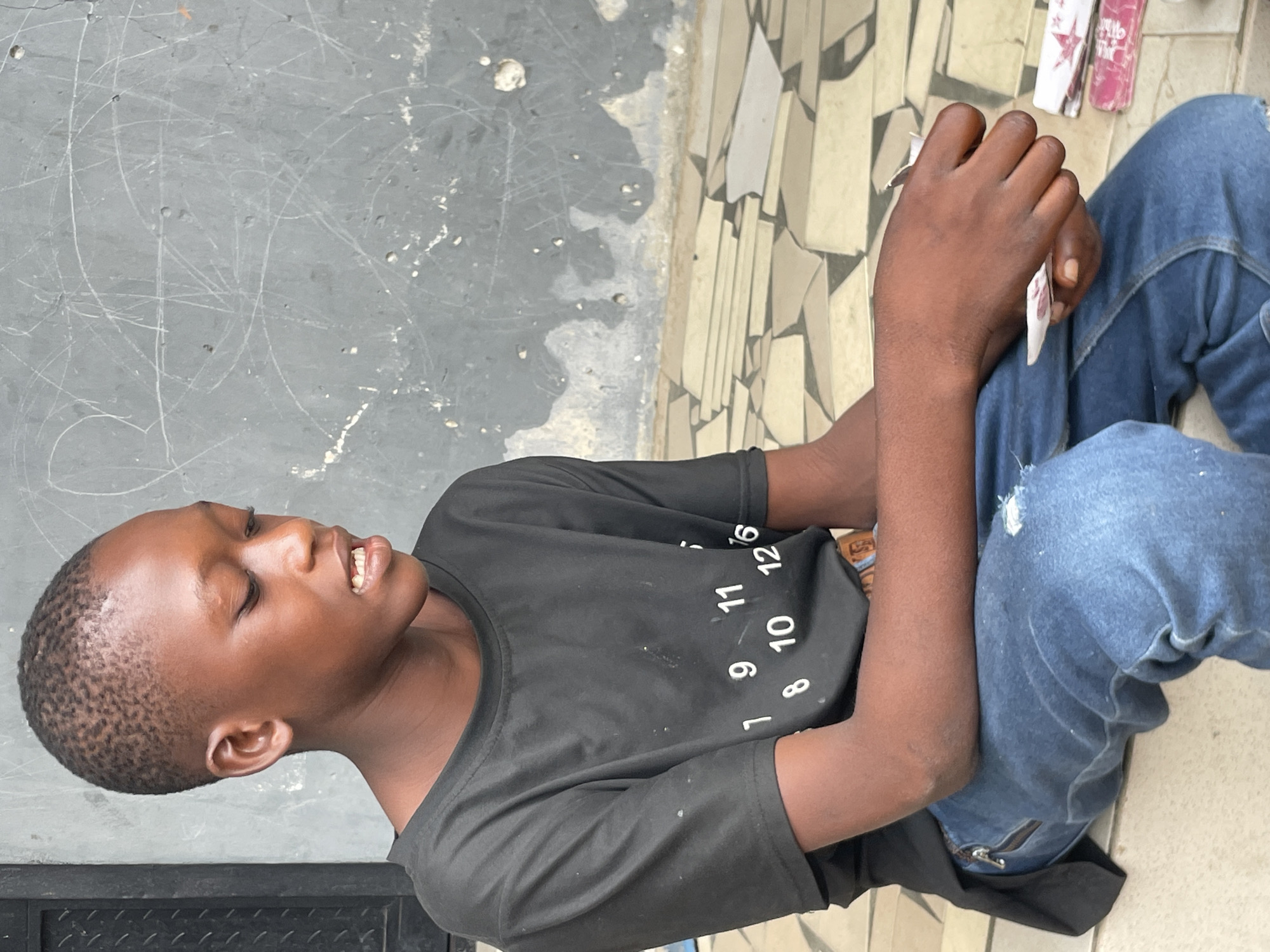 A young person, likely a boy, with dark skin and a short buzz cut, is captured in a vertical, eye-level shot. He is sitting on a tiled ground, leaning back against a dark gray, scratched wall. His head is tilted upwards and to his left, with his mouth slightly open in what appears to be a smile or laugh, suggesting an engaged or happy emotion.
He is wearing a black short-sleeved t-shirt with white numbers scattered across the front, clearly showing '1', '8', '9', '10', '11', '12', and '16'. His lower body is clad in blue jeans that appear distressed or ripped in places, with a zipper detail visible near his right knee. His right hand is raised and holds several small, folded papers or cards.
The ground is covered with light-toned, irregularly shaped mosaic-like tiles. In the background, behind his right hand, two small packets are visible: one pink with a white star and partially legible text that appears to include "Hair," and another white one. The bright, even lighting suggests it is daytime. This scene takes place in Badagry, Nigeria.