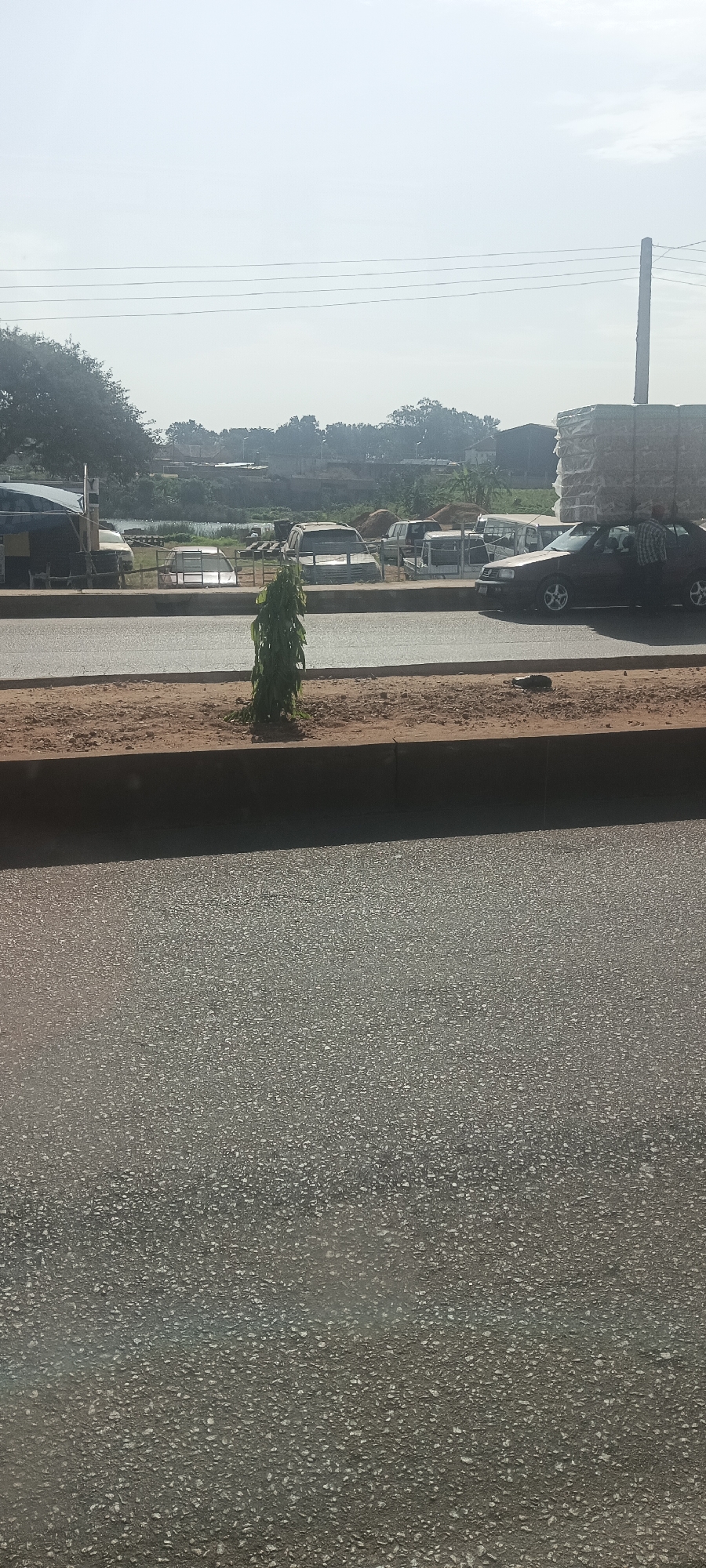 The image depicts a street in Bukuru, Nigeria, featuring an asphalt road with a raised median planted with a small, green tree. In the background, a row of parked vehicles is visible, including cars and trucks, some of which appear to be derelict or in storage. A man stands near a dark-colored car on the right, with what appears to be a load of materials stacked on the roof. Further back, a collection of low buildings and trees indicates the urban environment, with an overcast sky above. A utility pole is seen in the background.