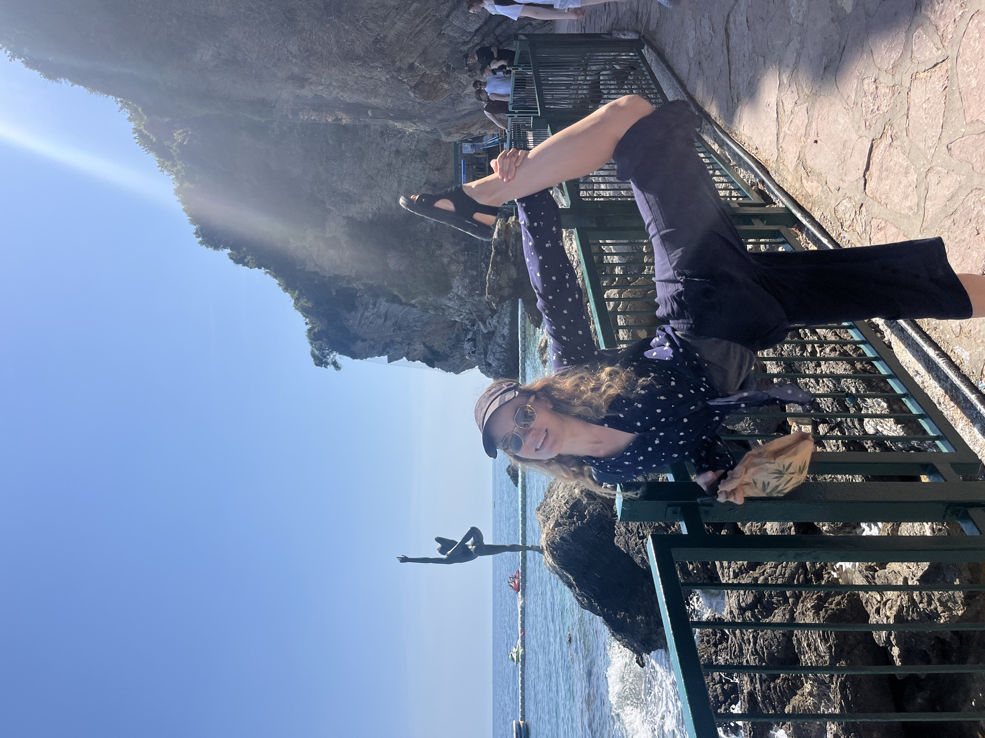 A woman is hanging on a green painted metal railing in Budva, Montenegro. The location is near the "Ballerina Statue" facing the Adriatic Sea. The woman is wearing a black cap, sunglasses, a navy polka dot shirt, and dark pants. She has one leg raised up, with a black sandal on her foot, over the railing. In the background, there are people on the cliff. There is also a rocky cliff side and the blue sky.