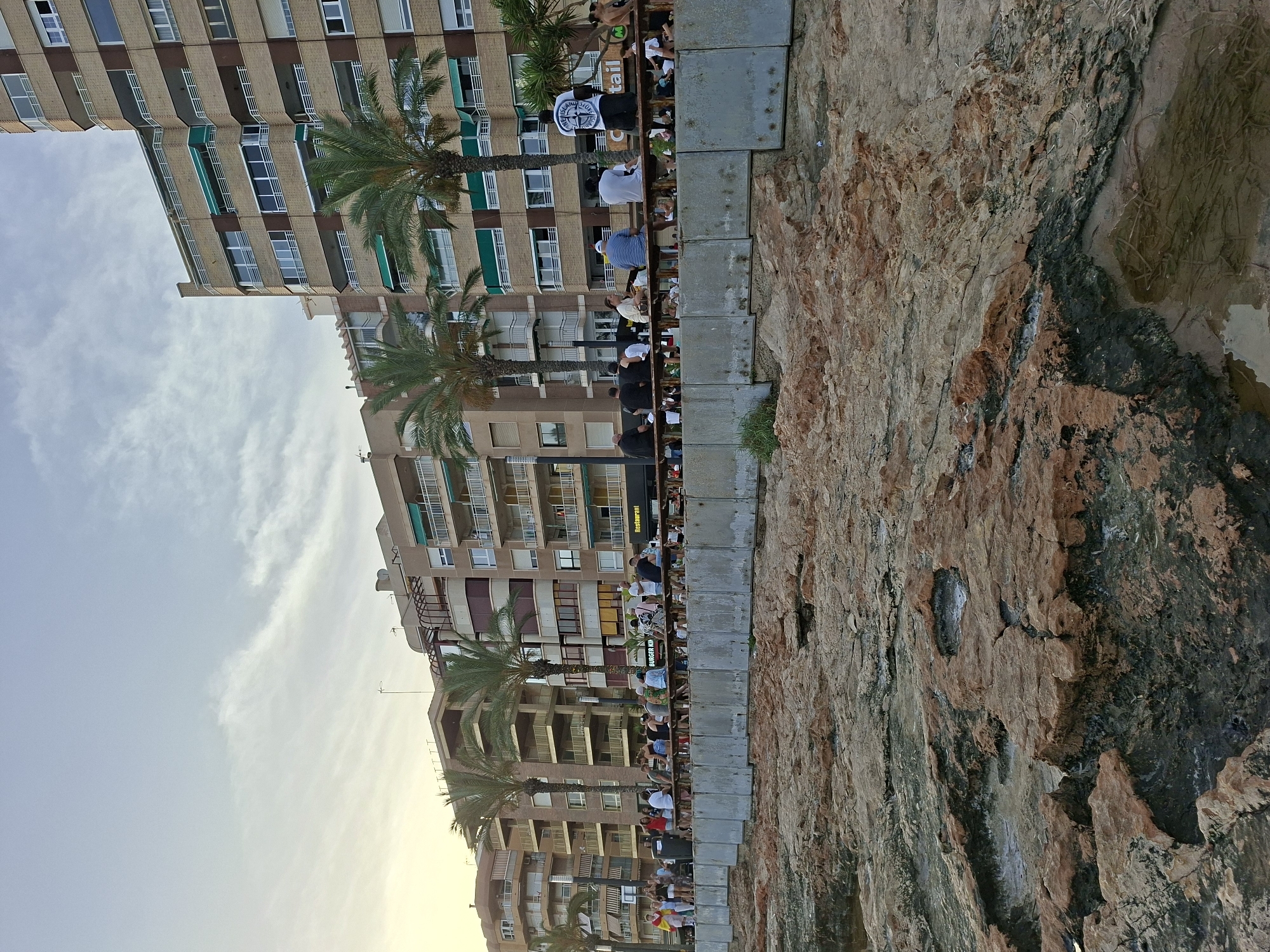 The image depicts a crowded promenade in Torrevieja, Spain, with a rocky shoreline in the foreground. A group of people are gathered along a railing, some seated and others standing. Behind them, several multi-story buildings with balconies and awnings line the promenade. Palm trees are interspersed between the buildings. The sky is partly cloudy. The rocky foreground shows patches of water and vegetation. The overall scene suggests a lively outdoor gathering or event.