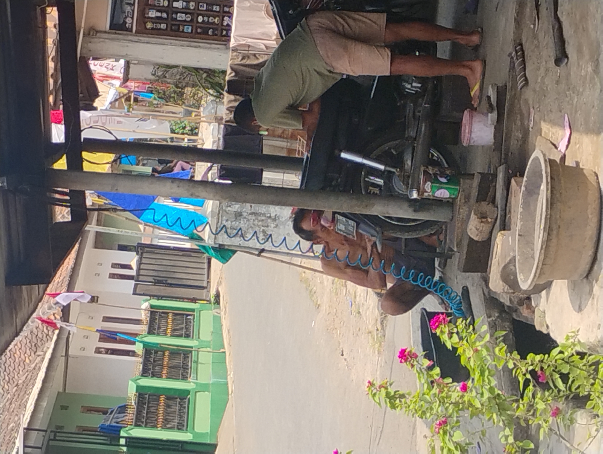 The image depicts an outdoor scene, likely a street or roadside repair area, in Nanggerang, Indonesia. Two men are present. One man, shirtless and wearing dark shorts, is seated on the ground, appearing to be working on a black motorcycle parked in the foreground. A blue coiled hose is draped around him and connected to the motorcycle. The second man, wearing a green t-shirt and light-colored shorts, stands near the front of the motorcycle, leaning over it and also appearing to be engaged in some activity related to the vehicle.
The environment includes a paved surface, possibly a street, and a dirt or gravel area where the men and motorcycle are situated. Various objects are scattered around, including wooden blocks, buckets, and what appears to be a tire. In the background, buildings with tiled roofs are visible, along with fences and flags, one of which is red and white, suggesting Indonesian national colors. Green painted structures and more distant houses are also present. A flowering plant with pink blossoms is in the lower left corner of the frame. The scene is illuminated by daylight, casting shadows.