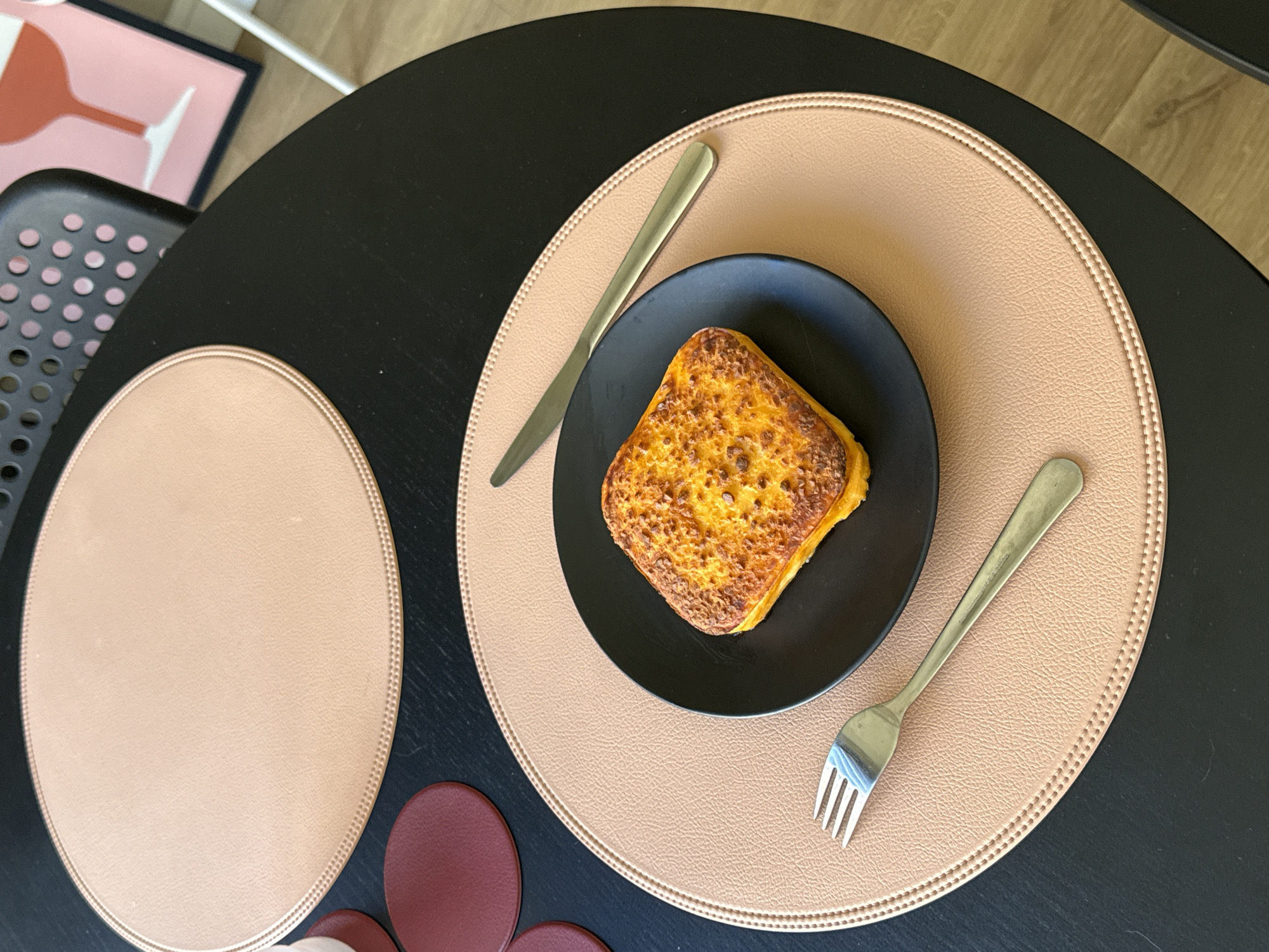 The scene depicts a black, round table with two beige placemats. A black plate with a golden-brown toasted sandwich sits atop one of the placemats, accompanied by a knife and fork. Under the second placemat, there are several dark red coasters. A black chair and a framed print in shades of pink are visible in the periphery. The location is indeterminate and there is no evidence to suggest it is in Barcelona, Spain. No people are present in the image.