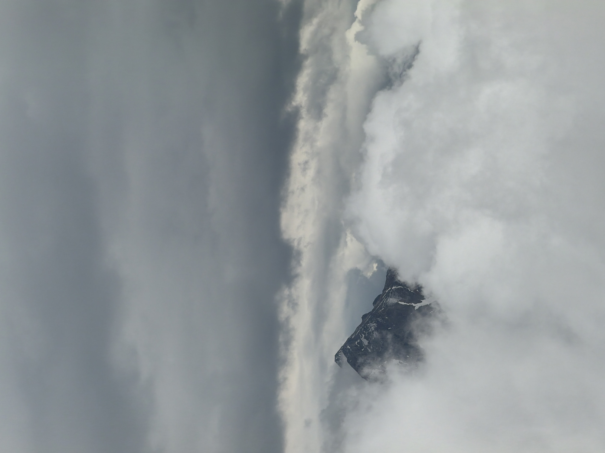 The image depicts a solitary, dark mountain peak shrouded in a dense, multi-layered cloud system under an overcast sky. The upper third of the frame is filled with a vast expanse of heavy, dark grey clouds, indicating deep atmospheric coverage. Below this, a lighter, more diffused band of grey clouds extends horizontally. Emerging from these middle layers of clouds is a rugged, sharp-peaked mountain, predominantly dark grey and brown in color, with discernible patches of white snow or ice clinging to its steep slopes and crevices. A smaller, secondary peak is partially visible behind and to the left of the main peak, also partially obscured by clouds. The lower half of the image is completely enveloped in thick, fluffy white and light grey clouds, obscuring the base of the mountain and creating a sense of height and isolation. The overall lighting is soft and diffused, characteristic of a cloudy day, with no direct sunlight. No human figures, animals, or man-made objects are visible. The scene is set in an alpine or mountainous region, location Unknown, Unknown.