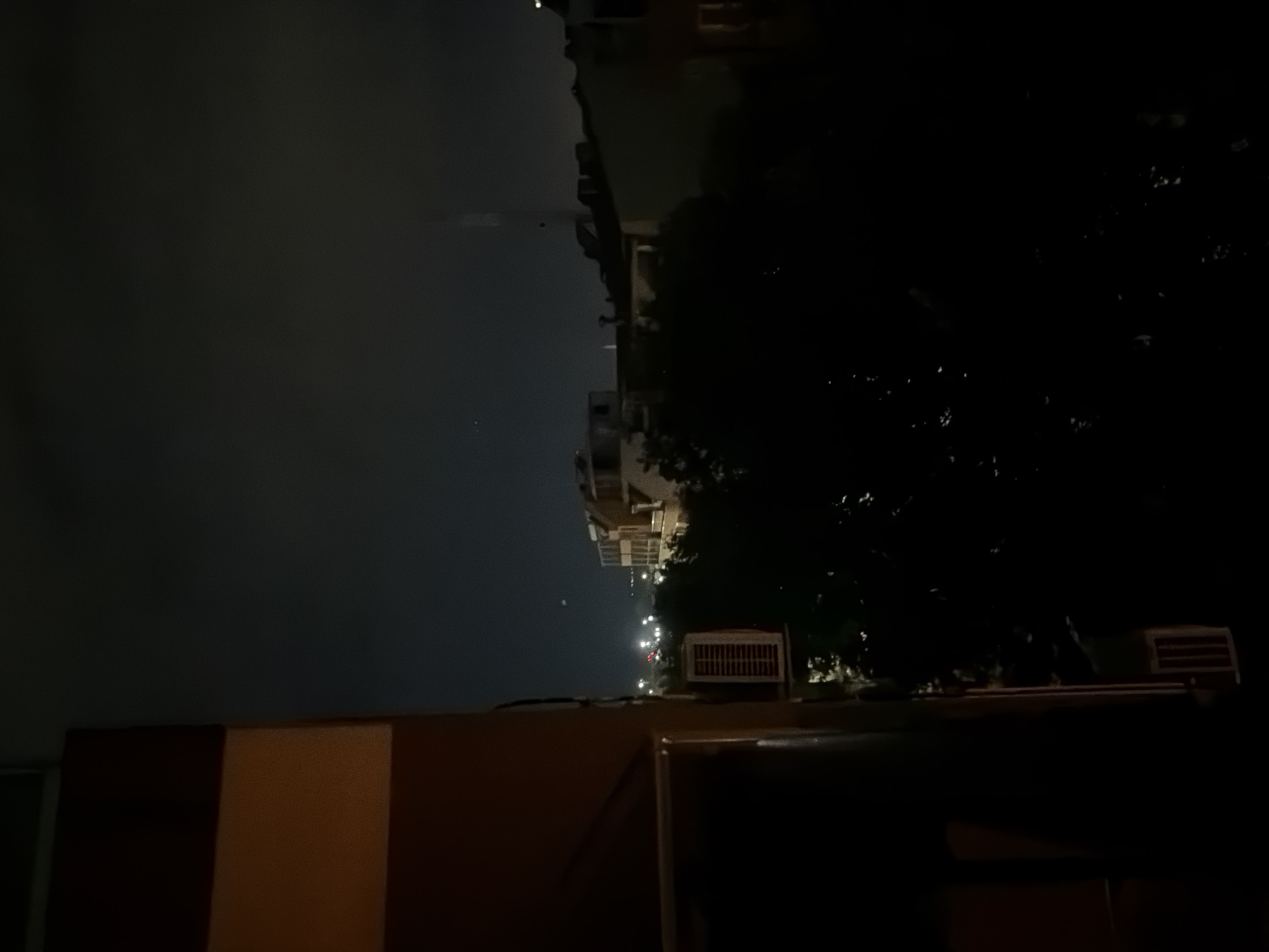 The image depicts a nighttime view from a building's balcony in Burgas, Bulgaria. The foreground is dominated by the dark side of a building. Beyond this, a city skyline is visible under a dark, cloudy sky. Buildings with illuminated windows and streetlights create a warm glow in the distance. Trees partially obscure the view of the city. An air conditioning unit is mounted on the exterior wall of the building in the mid-ground. The overall scene is dark and atmospheric, characteristic of a city at night.
