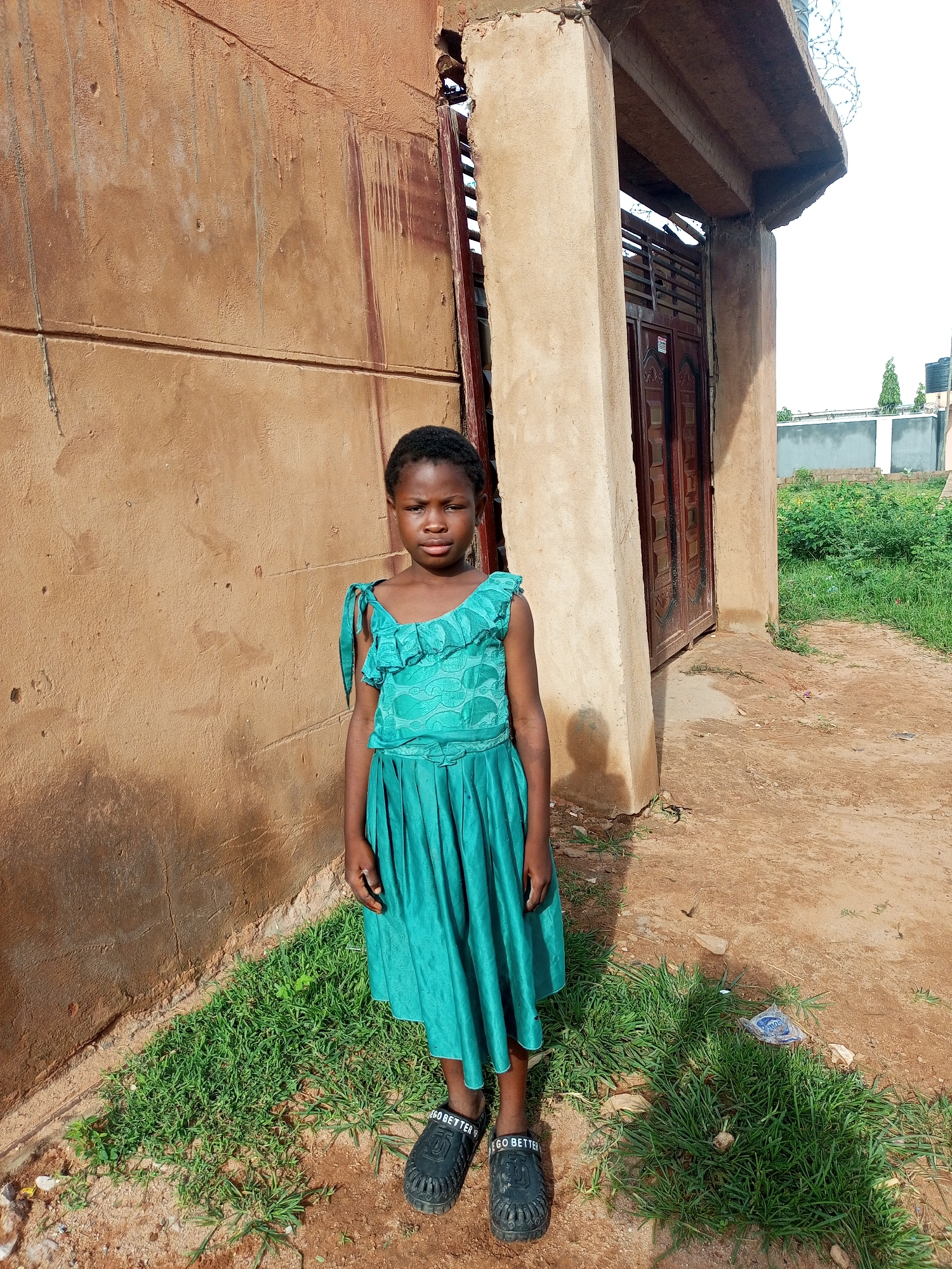 A young Black girl stands in front of a weathered, tan stucco wall in an outdoor setting. She is wearing a bright teal, sleeveless dress with ruffled accents around the neckline and shoulder straps. The dress has a fitted bodice and a full, pleated skirt. She is also wearing black slip-on shoes with white lettering that reads "GO BETTER." Her dark hair is cut short. The wall behind her shows signs of age and staining, with streaks of rust-colored water running down its surface. To the right of the girl, there is an entrance with a wooden double door set within a concrete archway, topped with a rough concrete overhang. Barbed wire is visible along the edge of the overhang. The ground in front of the wall is a mixture of dry, reddish-brown dirt and patches of green grass. In the background, beyond the entrance, lush green vegetation and some buildings are visible under a bright, overcast sky.