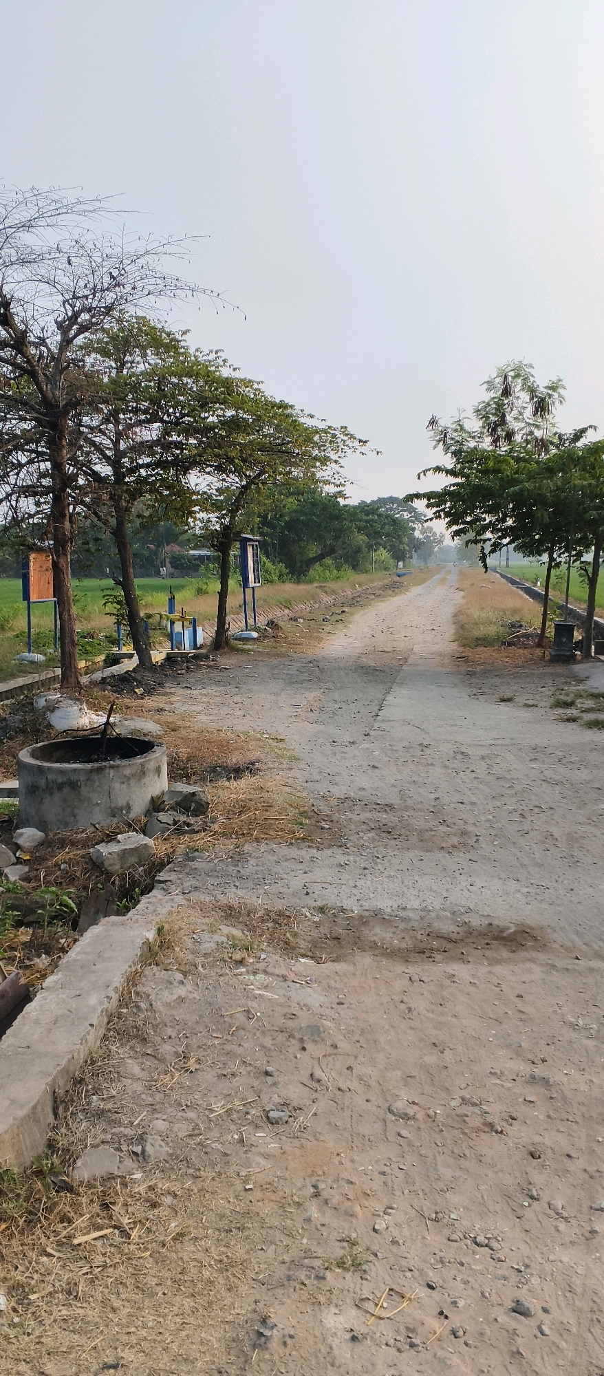 The image depicts a dirt road in a rural area, likely in Japanan, Indonesia, based on the context provided. The road appears to be a path leading through a landscape characterized by vegetation and some man-made structures.
On the left side of the road, there is a circular concrete well, which seems to be old and unused. Adjacent to the well, there are two trees with bare branches and some green foliage at the top. One tree has a wooden signpost attached to it. Further down the road on the left, there are two blue information boards mounted on poles, and a blue and yellow water control gate structure. Beyond these, a green field is visible, suggesting agricultural land. A narrow concrete channel runs alongside the road on the left.
The road itself is unpaved and dusty, with patches of dry grass and some scattered stones. In the distance, the road curves and disappears behind a line of trees. On the right side of the road, there is a row of trees with green leaves, and further down, another signpost and a small structure that could be a trash bin. The sky is overcast, indicating a possibly hazy or cloudy day. The overall impression is one of a quiet, somewhat rustic, rural setting.