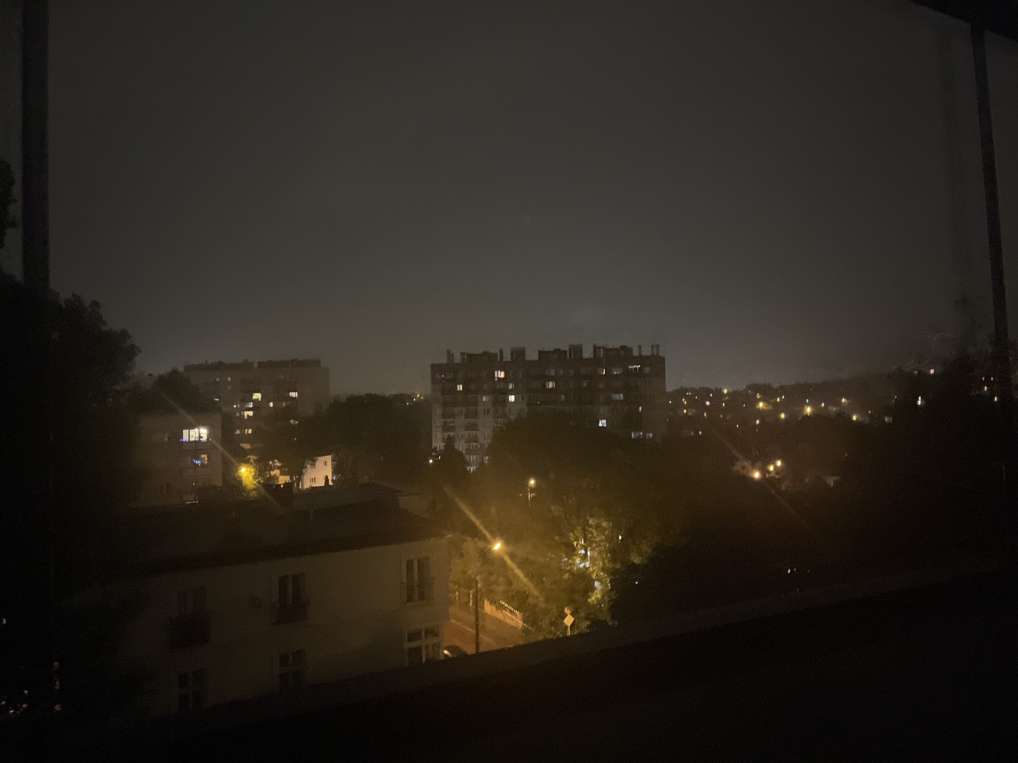 The image depicts a city skyline at night, shrouded in darkness and haze. The scene is dominated by the silhouette of several buildings of varying heights. In the foreground, a low-rise building with illuminated windows is visible. Further back, a large, multi-story apartment complex with many lit windows stands out. Trees, dark and indistinct, are scattered throughout the landscape, and streetlights cast a yellowish glow on the scene. The sky is a dark, murky gray, suggesting cloudy or foggy conditions. The overall impression is one of an urban environment at rest, with the lights of the buildings piercing the nighttime gloom.
