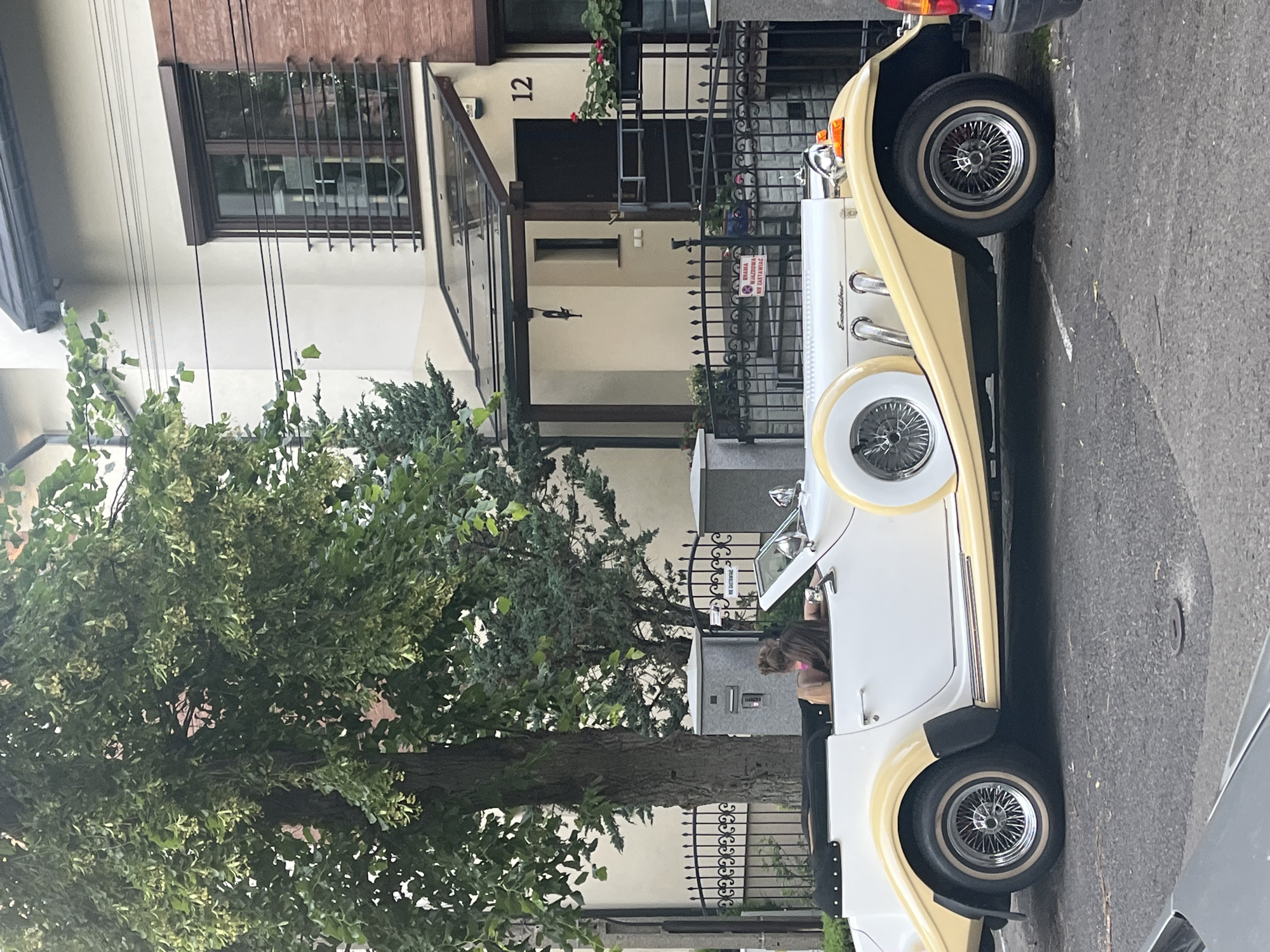 A side profile of a cream and yellow vintage convertible is parked on an asphalt street in front of a building. A woman with long brown hair, wearing a pink top, is seated in the driver's seat of the car, facing away from the camera. The car features chrome accents, wire wheels, and a spare tire mounted on the side. Behind the car, a modern house with a light-colored facade, dark-trimmed windows, and a glass-covered entryway is visible. A black metal fence with decorative elements separates the property from the street. Trees with green foliage are present on the left side of the image. The sky is overcast. A street sign with the number "12" is displayed on the building. A smaller, dark-colored vehicle is partially visible to the right of the vintage car.