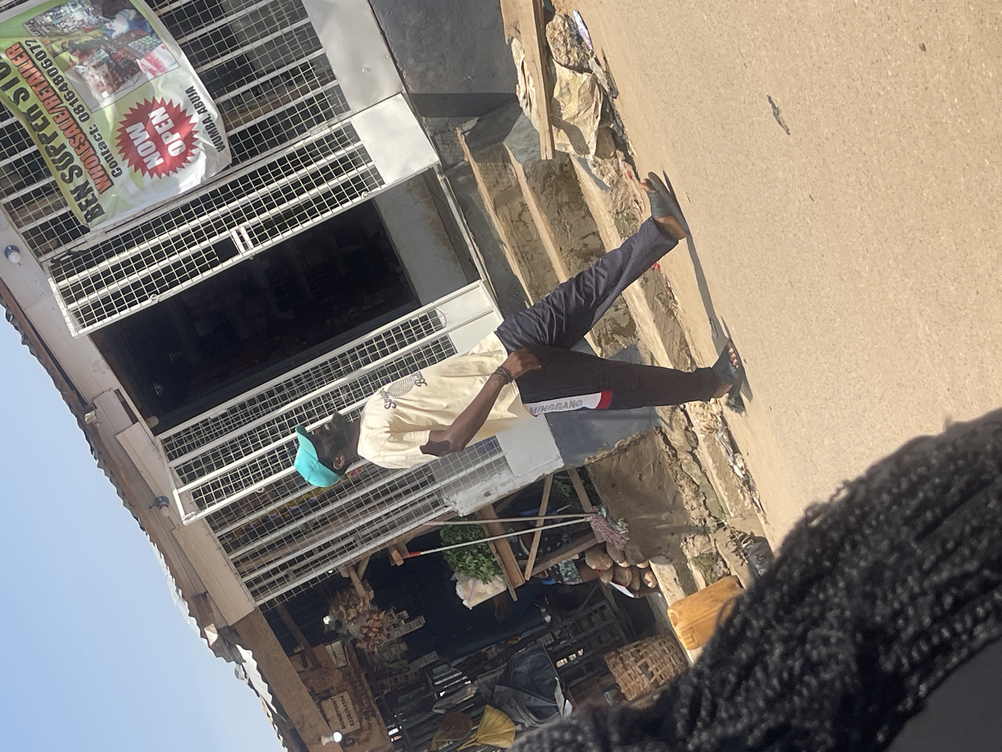 A man in a light-colored t-shirt and dark pants, wearing a teal baseball cap and black flip-flops, stands on a street in Wumba, Abuja, Nigeria. He is facing to his right, with his body angled away from the viewer. Behind him is a storefront with a metal gate and a banner that reads "BEN SUPER STORE WHOLESALE/RETAILER Contact: 08164806072 NOW OPEN WUMBA, ABUJA". The store appears to be open, with goods visible inside. The man is walking or posing on the paved street in front of the store. The scene is illuminated by bright sunlight, casting shadows on the ground. In the foreground, the braided hair of a person is visible, obscuring part of the scene. Various objects are scattered on the ground, including a yellow plastic container and baskets. The overall impression is of a busy, outdoor commercial area.