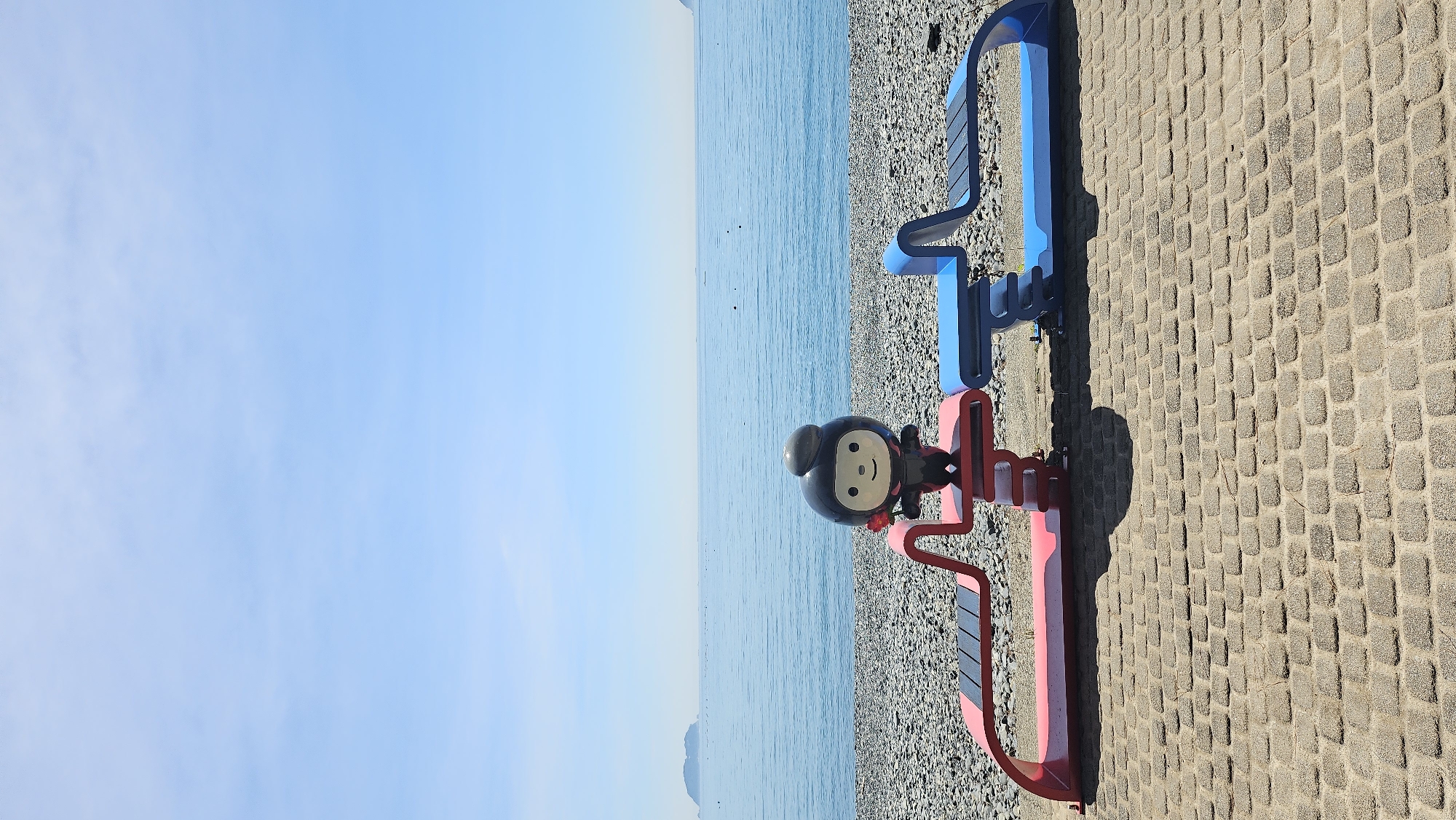A cartoon-like mascot stands on a red and blue abstract sculpture facing a calm, blue ocean under a clear sky. The mascot is black with a round head, a smile, and a small grey hat. It holds a red flower in its right hand. The sculpture is situated on a paved walkway, with a pebbled beach and the sea stretching to the horizon. In the distance, a small island is visible on the left, and another landmass on the right. The scene is likely located in Geoje-si, South Korea, given the prevalence of coastal areas and the distinct aesthetic of public art installations in the region.