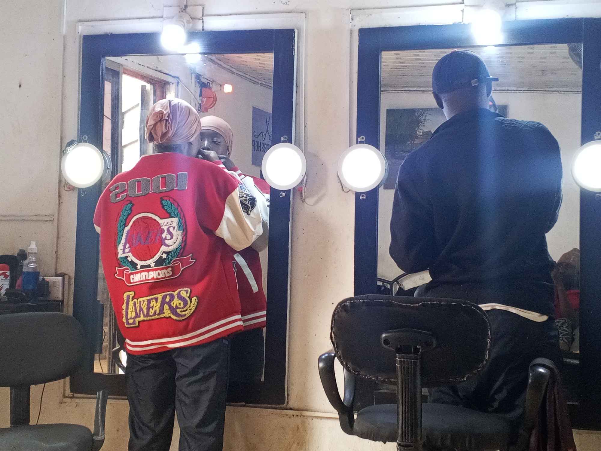 The photo depicts the interior of what appears to be a small barbershop in Gwong, Nigeria. There are two barber chairs, one in the foreground and one to the left. A person wearing a red Lakers champion jacket and a head covering is standing in front of a mirror, apparently being attended to by another person who is reflected in the mirror. Another person is sitting in the barber chair to the right, looking at a mirror. The mirrors are framed and have lights attached to them. There are some hair care products visible on a shelf behind the person in the Lakers jacket. The overall condition of the space suggests a modest, well-used establishment.
