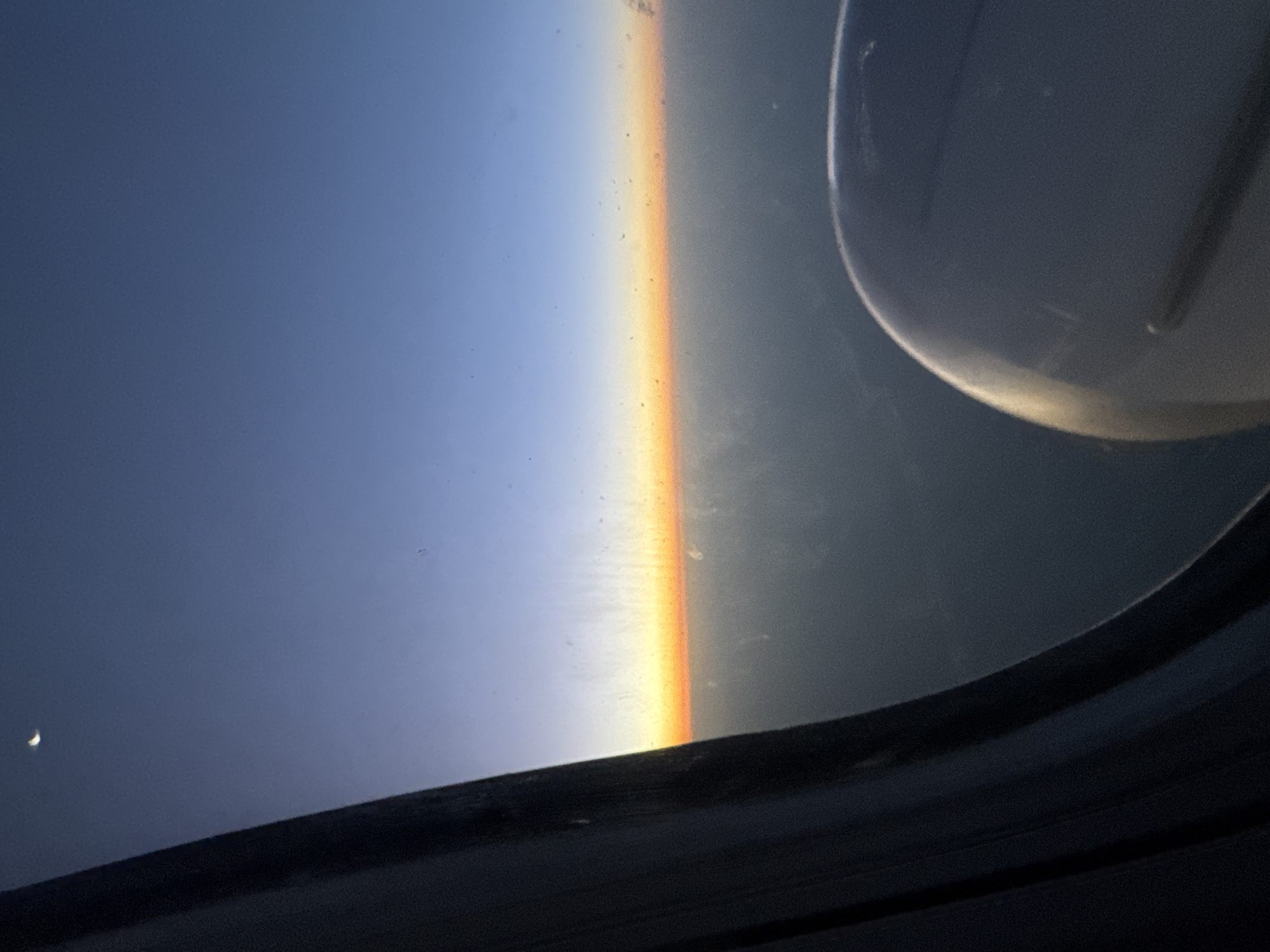 The image depicts a view from an airplane window during what appears to be either sunrise or sunset. The sky transitions from a deep blue at the top to a pale orange horizon line. A sliver of a crescent moon is visible in the upper left corner. A dark, textured curtain lines the left edge of the frame, obscuring part of the view. In the lower right corner, a portion of an airplane engine is visible. The scene is set above a layer of clouds or haze, with the landscape below indistinct. The image is grainy, suggesting it was taken in low light conditions or with a mobile device. The provided crops offer closer views of the moon, the sky, the horizon, and the airplane engine. Based on the provided information, this image is not specifically located in Monoq, Uzbekistan, as there is no information within the image or its crops to suggest this location.