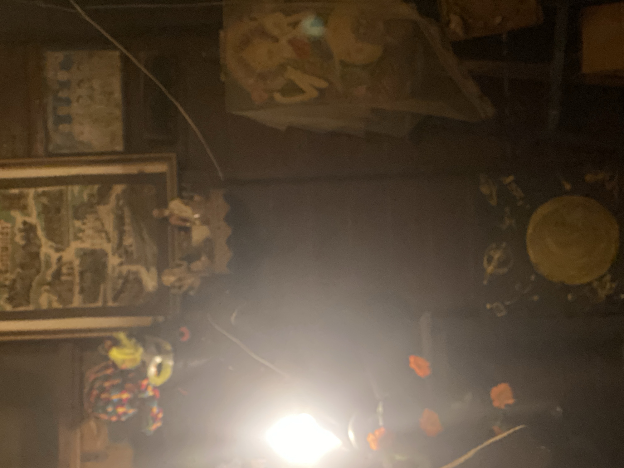 The image, potentially taken in Baan Don Sai, Thailand, shows a cluttered interior space. Hanging on a dark wall are two framed images, one above the other on the left. Further right, an indistinct plastic bag containing various items hangs from the ceiling. A decorative plate is mounted on the wall in the upper right corner. Below the plate are faint, blurry images of orange flowers. A light source obscures the bottom of the frame.