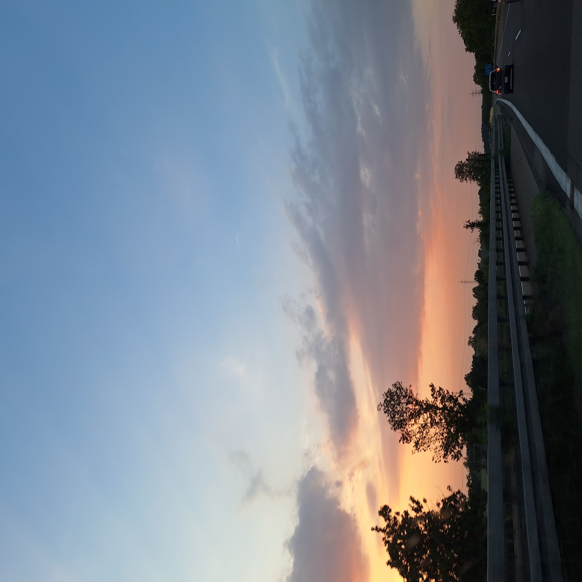The image depicts a highway scene at dusk. The sky is a gradient of pale blue at the top, transitioning to shades of pink and orange near the horizon, indicating sunset. Dark, silhouetted trees line the sides of the highway, with a few scattered power line poles visible in the distance. A metal guardrail runs along the left side of the road. On the right side of the highway, a dark-colored car with its brake lights illuminated is driving away from the viewer. The road appears to be paved, with white lane markings visible. The overall impression is of a travel scene during twilight.