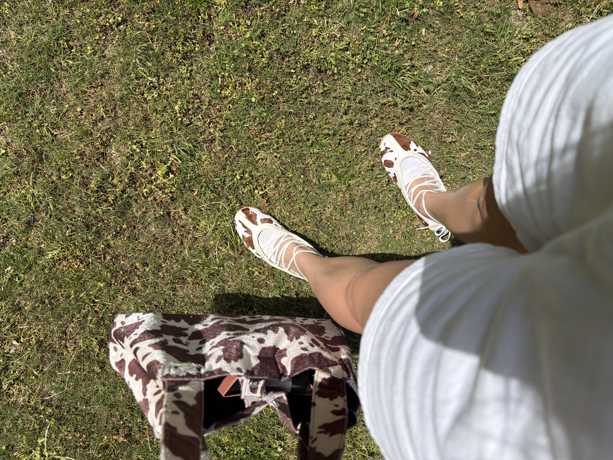 A medium, eye-level shot depicts an individual standing on a grassy lawn, possibly within a park or garden. The person is wearing a white, pleated garment and cow-print shoes with white laces. A cow-print tote bag is positioned near their feet. The environment is sunny, with shadows cast across the grass. While the location is not definitively discernible from the image alone, the prompt specifies Tel-Aviv, Israel.