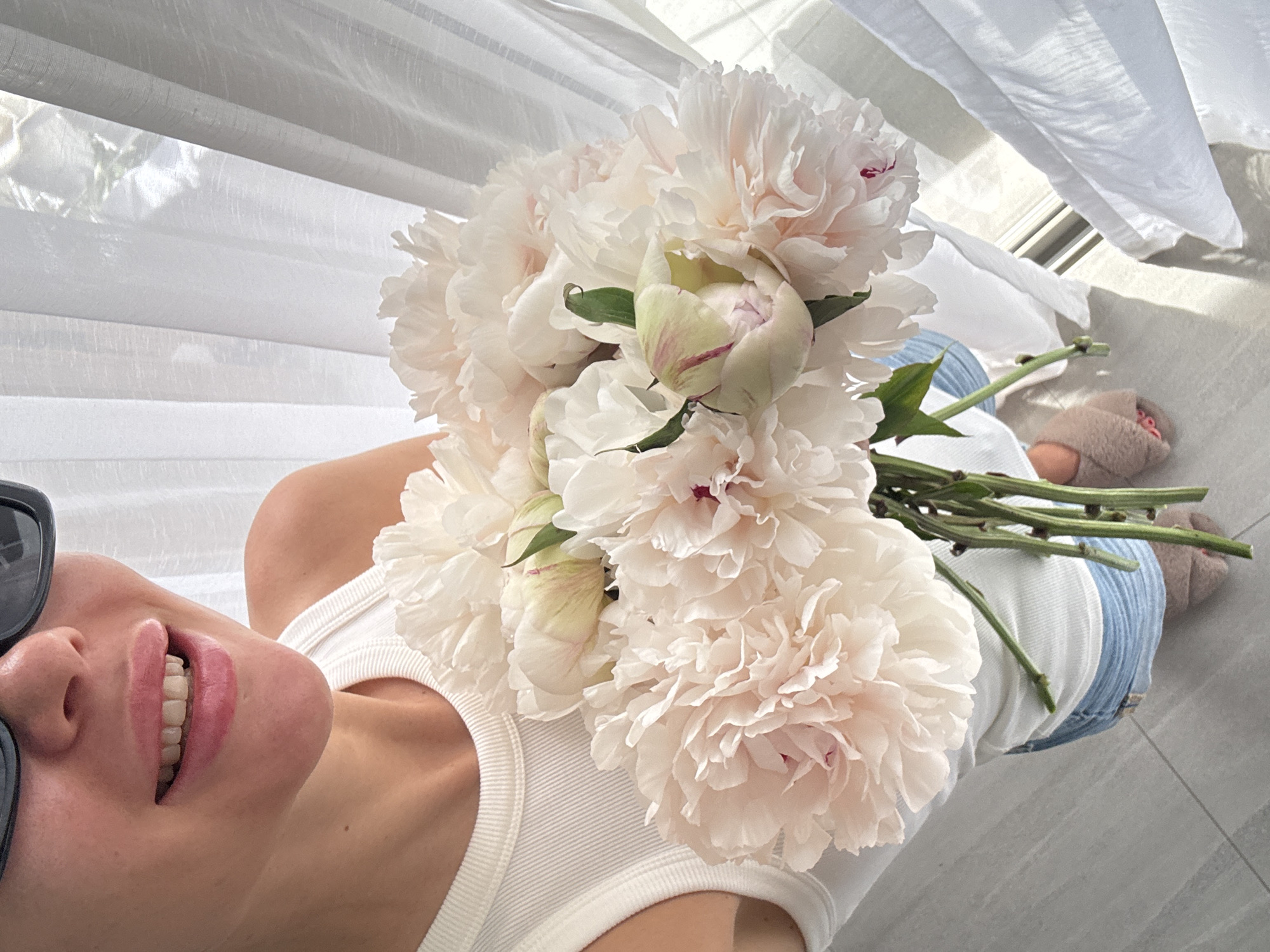 In Choeng Thale, Thailand, a partial upper body shot captures a person from the chest down, holding a large bouquet of approximately nine light pink and white peonies, some in bloom and some as buds, by their green stems. The person wears dark rectangular sunglasses, obscuring their eyes, and smiles, revealing their upper teeth and pink-glossed lips. They are dressed in a white ribbed tank top, light blue denim shorts, and a fluffy pink slipper is visible on their right foot. The background features light grey tiled flooring on the left and white sheer curtains on the right, through which bright natural light is visible, suggesting a window. The overall scene is brightly lit.