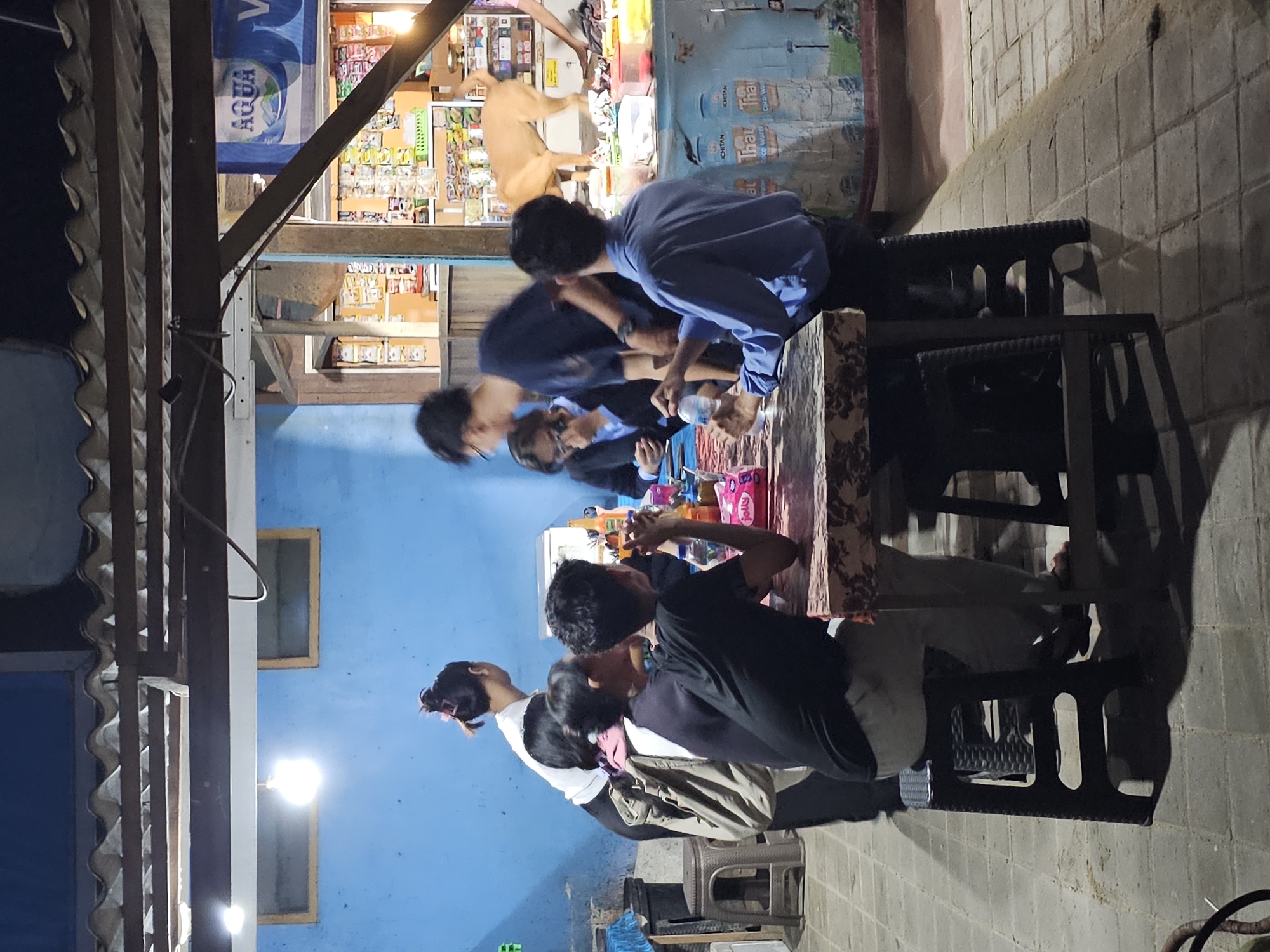 A group of about seven people are sitting around a table at an outdoor establishment in Denpasar, Indonesia, possibly a cafe or warung. The setting is at night. Some of the people are looking down at the table or their phones, while others are talking. They are seated on plastic chairs. The location appears to be next to a small shop selling various items. The building is painted blue.