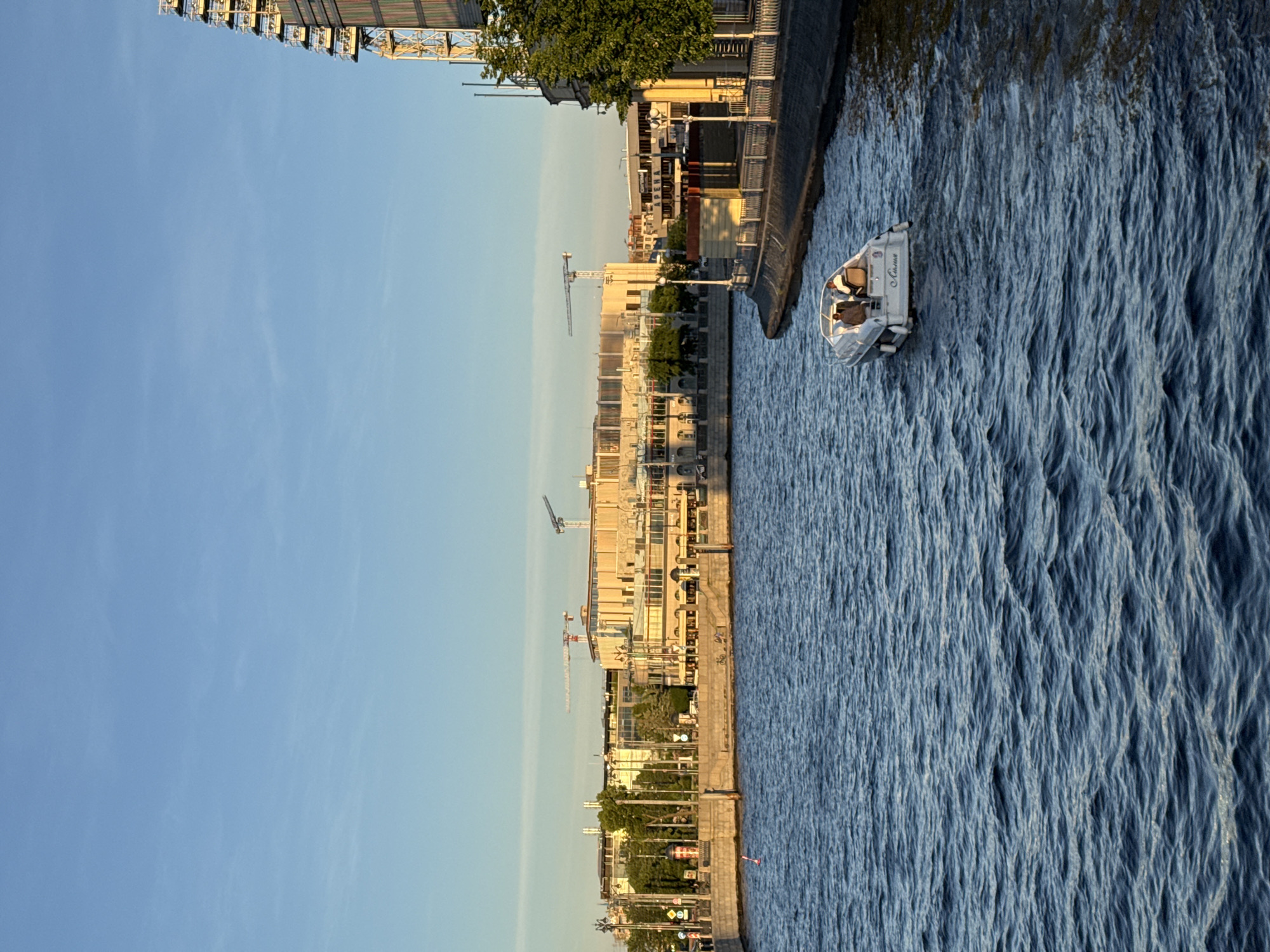 This is a horizontal, eye-level shot of a river in Saint Petersburg, Russia, at golden hour. The water is dark blue and choppy. A small white motorboat with two people aboard is moving away from the viewer on the right side of the frame. The boat has the name "Айвазовский" inscribed on its stern.
The riverbank on the right is a dark concrete wall with a narrow paved walkway and some trees. On the left bank, a long, light-colored building with a colonnade of windows is visible, with trees and streetlights in front of it. In the background, more buildings and construction cranes indicate ongoing development. The sky is clear blue with a few wispy clouds. A tall structure, possibly a Ferris wheel or observation tower, is partially visible on the far right edge of the frame.