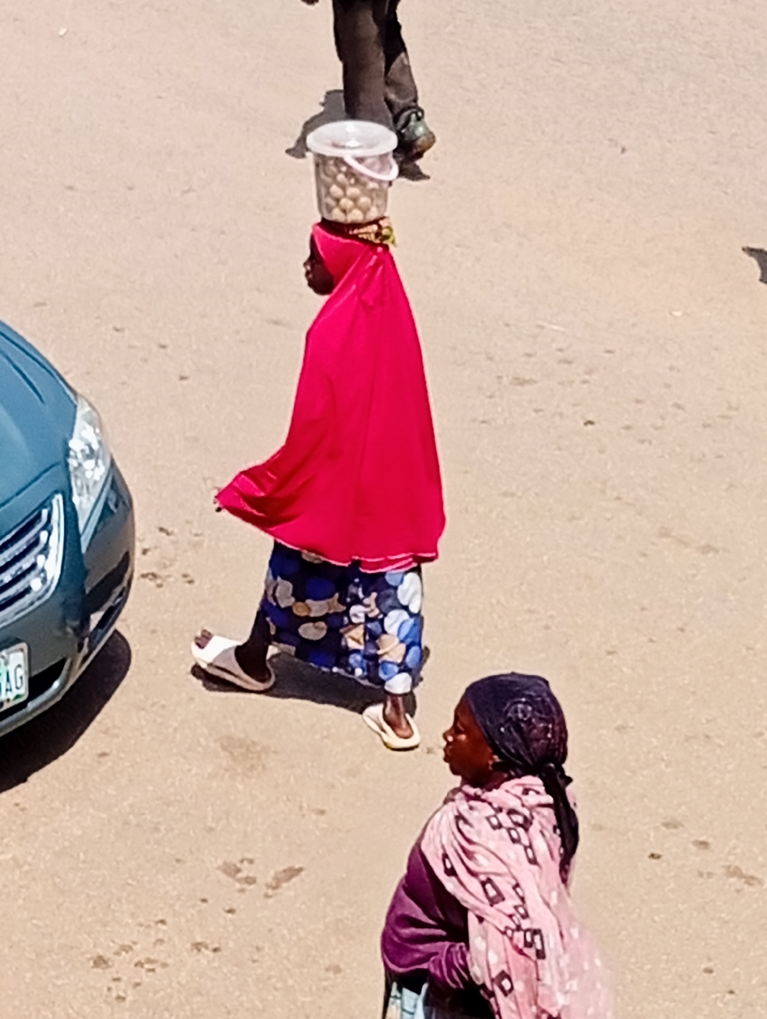 This image shows two women on a street in Bukuru, Nigeria. The woman in the foreground is wearing a dark headscarf and a pink patterned scarf over a purple garment. She is looking to the left, with her head tilted slightly.
Behind her and to her left, another woman is walking. She is wearing a bright pink headscarf and a blue and white patterned skirt, with a red garment draped over her shoulders. She is carrying a clear plastic bucket filled with round, light-colored objects on her head. The objects appear to be food items, possibly snacks or sweets. Her left foot is visible, wearing a white sandal.
A portion of a dark green car is visible on the left side of the image, with a license plate that appears to have "DAG" printed on it. The ground is a light-colored, sandy or dusty surface, possibly a road or a pathway. The lighting suggests it is daytime, likely with bright sunlight, as indicated by the shadows. The scene appears to be an outdoor setting on a street, typical of a marketplace or a busy thoroughfare in Nigeria.