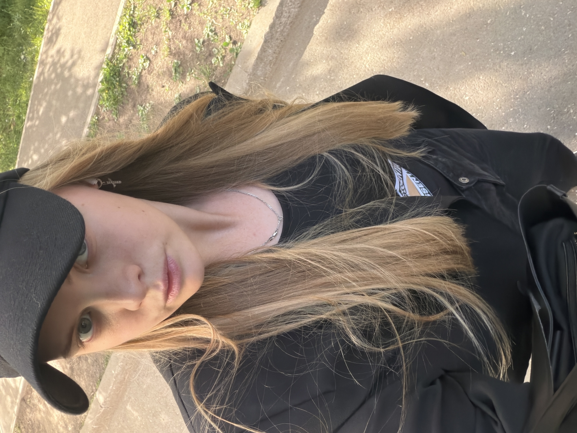 A woman with long, blonde hair is captured in a selfie, wearing a black baseball cap and a black jacket. She is facing the camera with a slight upward tilt. A silver chain necklace is visible around her neck. The background suggests an outdoor setting with greenery and a paved area, potentially a sidewalk. The specific location is not discernible.