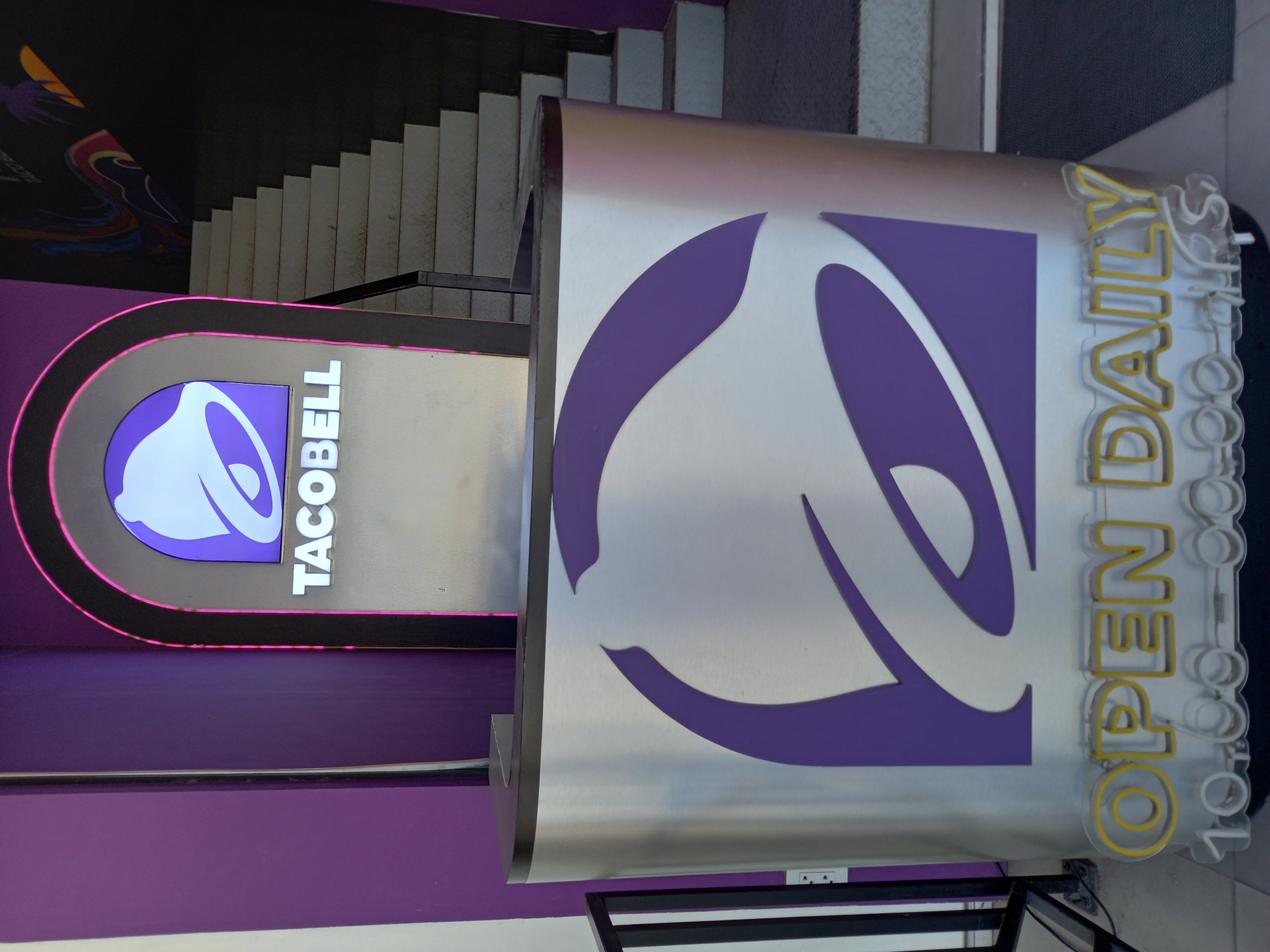 A Taco Bell restaurant with a modern, metallic exterior and purple accents is visible in the city of Baan Bo Phut, Thailand. The restaurant features a prominent illuminated Taco Bell logo and a sign reading "OPEN DAILY" with the hours "10:00 AM - 10:00 PM". The entrance is flanked by purple walls and a staircase is visible in the background. The scene is well-lit, suggesting it is daytime or the restaurant is brightly lit internally.