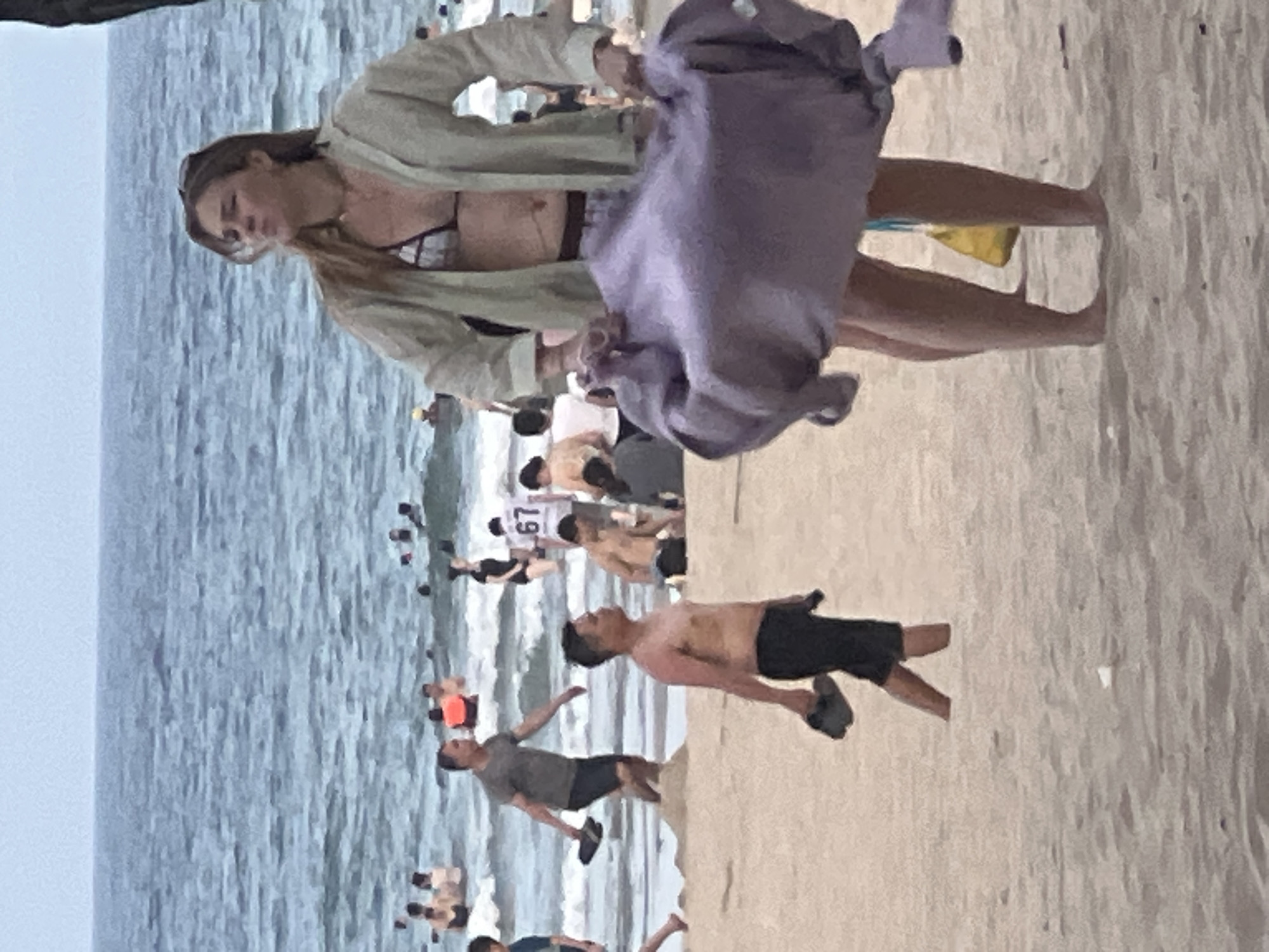 The image depicts a crowded beach scene at Đà Nẵng, Vietnam. The foreground is dominated by a person in the process of putting on or taking off a light-colored, loose-fitting jacket over a bikini. They are holding a purple garment, possibly a towel or another piece of clothing, in their hands.
In the mid-ground, the ocean is visible with gentle waves, and numerous people are scattered along the shoreline and in the water. Some individuals are wading or swimming, while others are standing and interacting. Further back, more people are visible on the beach, suggesting a busy day.
A man in the mid-right of the frame is captured in motion, appearing to be running or jumping. He is shirtless, wearing dark shorts, and has dark hair. Another man in the mid-left is also in motion, wearing a grey t-shirt and dark shorts.
There are signs of activity and recreation on the beach, with groups of people gathered. The lighting suggests it could be daytime, possibly overcast or late afternoon, given the muted colors and lack of harsh shadows. The overall atmosphere appears to be lively and social.
A notable detail visible is the number "67" printed on a white shirt worn by someone in the background among the crowd near the water. The setting is clearly a beach, with sand in the foreground and the ocean stretching out behind the people.