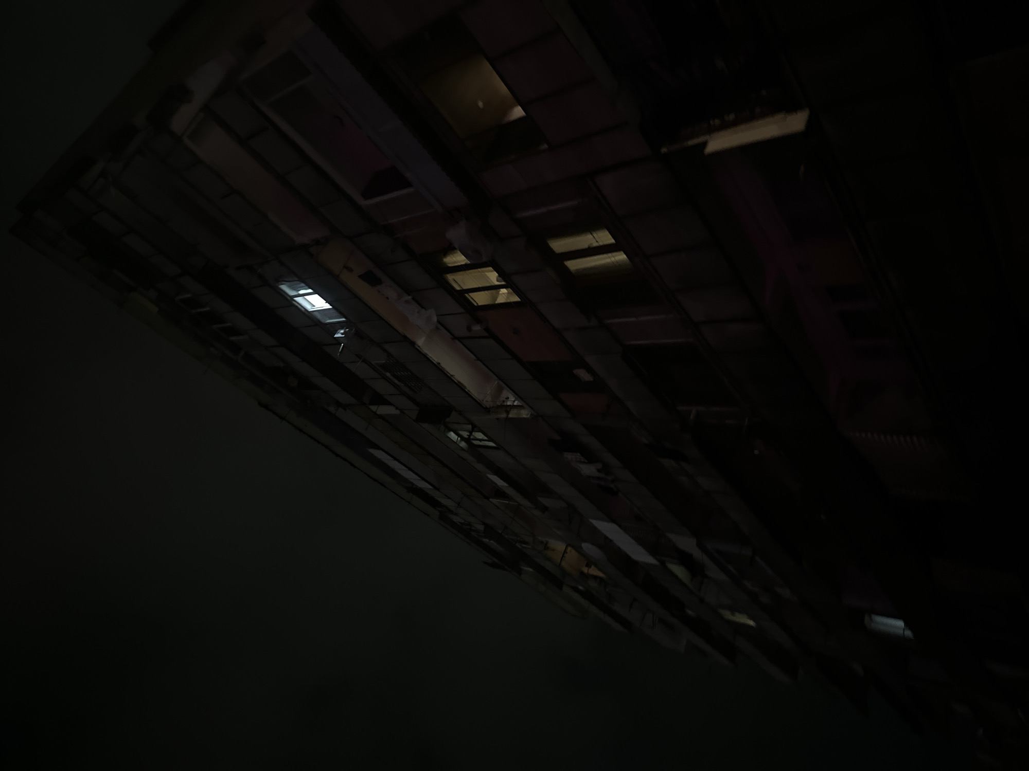 The image presents a dark, low-light view looking upwards at a large, angular structure. The perspective is tilted, making the structure appear to recede diagonally from the upper left to the lower right of the frame, with a vast expanse of darkness occupying the remainder of the image.
The primary subject is an overhead structure composed of numerous rectangular panels and beams, possibly part of a building's ceiling, an underpass, or a bridge. Several rectangular openings or fixtures within this structure emit faint, cool white or yellowish light, suggesting interior illumination or windows viewed at night. One particularly bright white rectangular light is prominent on the left side of the structure. Other fainter lights or reflections are visible across the panels. Hints of purple or pink hues can be subtly discerned in some of the darker, reflective surfaces on the right side.
No people or discernible activities are taking place. The scene conveys a sense of stillness and architectural scale. The overwhelming darkness indicates either nighttime or a very dimly lit indoor/covered environment. There is no visible text in the image. The unique upward and angled composition creates an abstract, somewhat disorienting visual experience.
