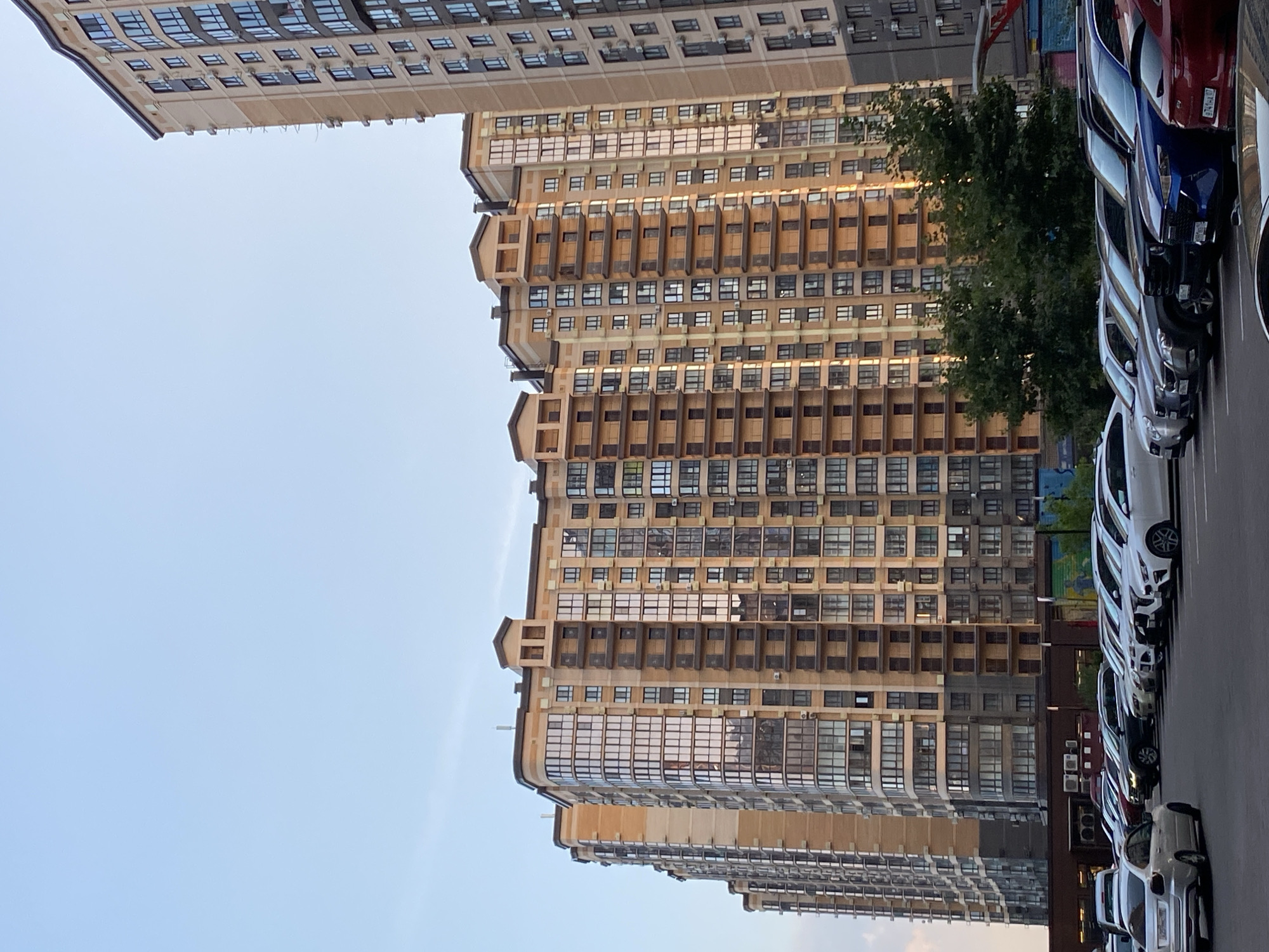 The image shows a parking lot in front of several tall residential buildings. The buildings are modern, with many windows and balconies, and are constructed with a combination of beige and brown materials. The parking lot is filled with a variety of cars, including sedans and SUVs. The sky is clear and blue, suggesting it is daytime. Some trees are visible in front of the buildings. The overall scene depicts a typical urban residential area.