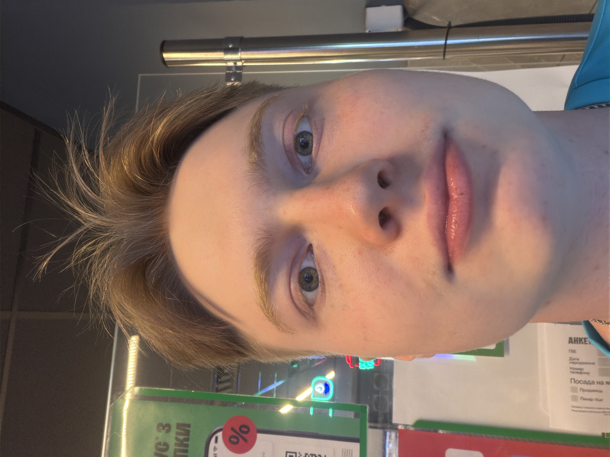 A close-up, selfie-style photograph features a young person with light brown, spiky hair. The subject's face is the primary focus, with their eyes open and looking towards the camera. Their skin appears fair with some freckles and slight redness. A portion of a green sign with text in Cyrillic and a red circle with a '%' symbol is visible on the left side of the image. Behind the subject, a metal pole is present, and the background appears to be an indoor setting with some blurred lights and signage. The image is taken from a low angle, looking up at the subject. There is no discernible location in the city of Kyiv, Ukraine, that can be identified from the visual information provided.