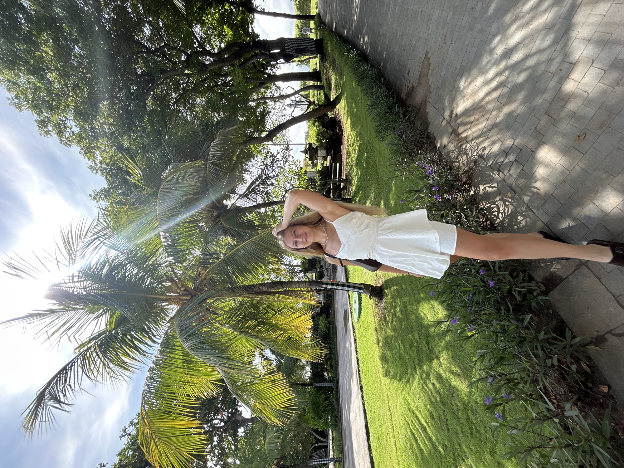 A young woman is pictured in a bright, tropical outdoor setting, consistent with Kuta Selatan, Indonesia. She stands on a well-maintained green lawn beside a paved stone pathway, looking towards the camera with a relaxed smile, one hand casually resting on the back of her head. She has long blonde hair and is wearing a white dress and dark shoes.
The environment is lush with vegetation, dominated by several tall palm trees, some bearing coconuts, whose fronds are illuminated by intense sunlight, creating noticeable lens flares. Other dense green trees and foliage are visible, and in the foreground, small bushes with delicate purple flowers line the edge of the path. The pathway itself is made of light-colored stone pavers, upon which strong, intricate shadows of leaves are cast by the overhead sun. In the background, deeper within the foliage, some tree trunks are adorned with a distinctive black and white checkered cloth, common in Balinese culture. Beyond the trees, a glimpse of what appears to be a body of water or distant landscape can be seen under a bright, partly cloudy sky. The bright light suggests it is daytime. There is no visible text.