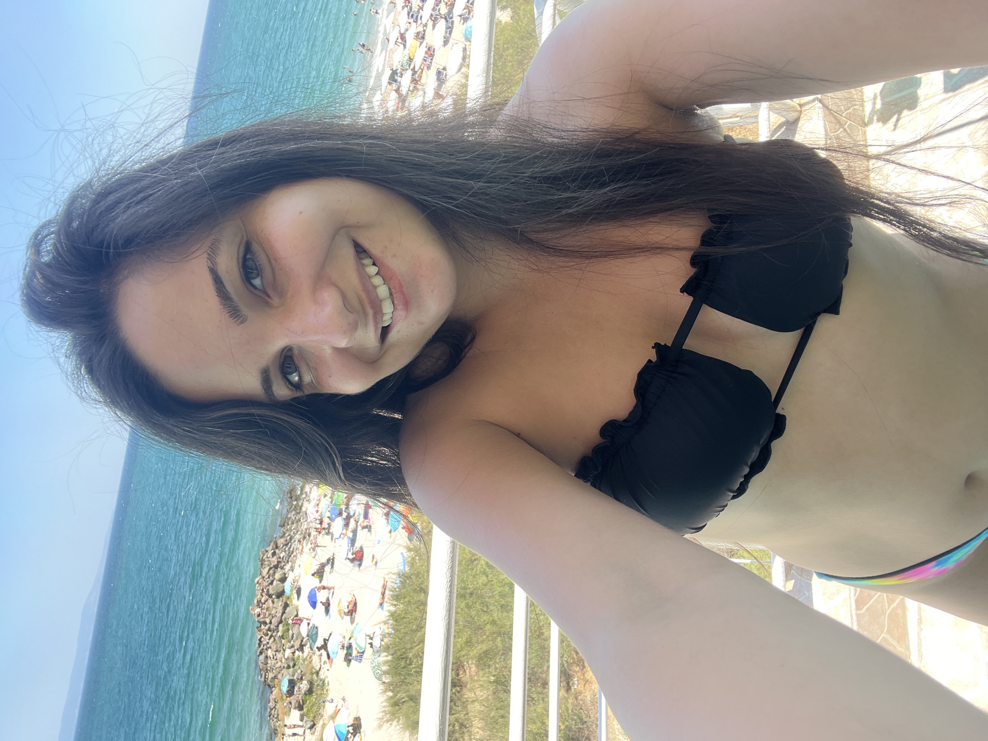 A young woman with long, dark hair and a bright smile is taking a selfie on a balcony overlooking a beach in Pomorie, Bulgaria. The woman is wearing a black bikini top and colorful bottoms. The beach is populated with many beachgoers relaxing under umbrellas. The sea is a bright blue, and the sky is clear. The balcony railing is white, and there are some trees and shrubs visible in the background. The image is taken in daylight.