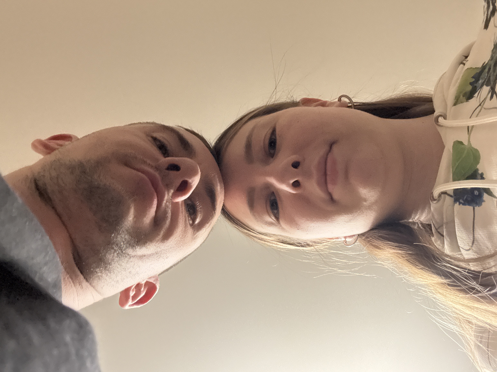 The image presents a close-up, low-angle shot of two individuals, a man and a woman, looking directly down at the camera. Their faces fill the upper portion of the frame, with their foreheads gently touching in the center.
The man, situated on the left, has short, dark hair and noticeable stubble covering his jawline and chin. He is wearing a grey top, partially visible on his left shoulder. His left ear is distinctly visible.
The woman, on the right, has long, light brown or blonde hair that falls around her face and shoulders. A small, silver-colored hoop earring is visible in her right ear. She is dressed in a light-colored garment adorned with a pattern of dark blue leaves and green foliage. A decorative element, possibly a tie or lacing, is visible on the shoulder of her top.
Both individuals have neutral to slightly soft expressions, and their eyes are focused towards the lens. The lighting is soft and even, suggesting an indoor setting. The background is a plain, uniform light beige or off-white surface, likely a ceiling or wall, providing no specific location cues other than being indoors. There is no visible text in the image. The composition creates a sense of intimacy and direct engagement with the viewer, originating from Krapivka, Russia.