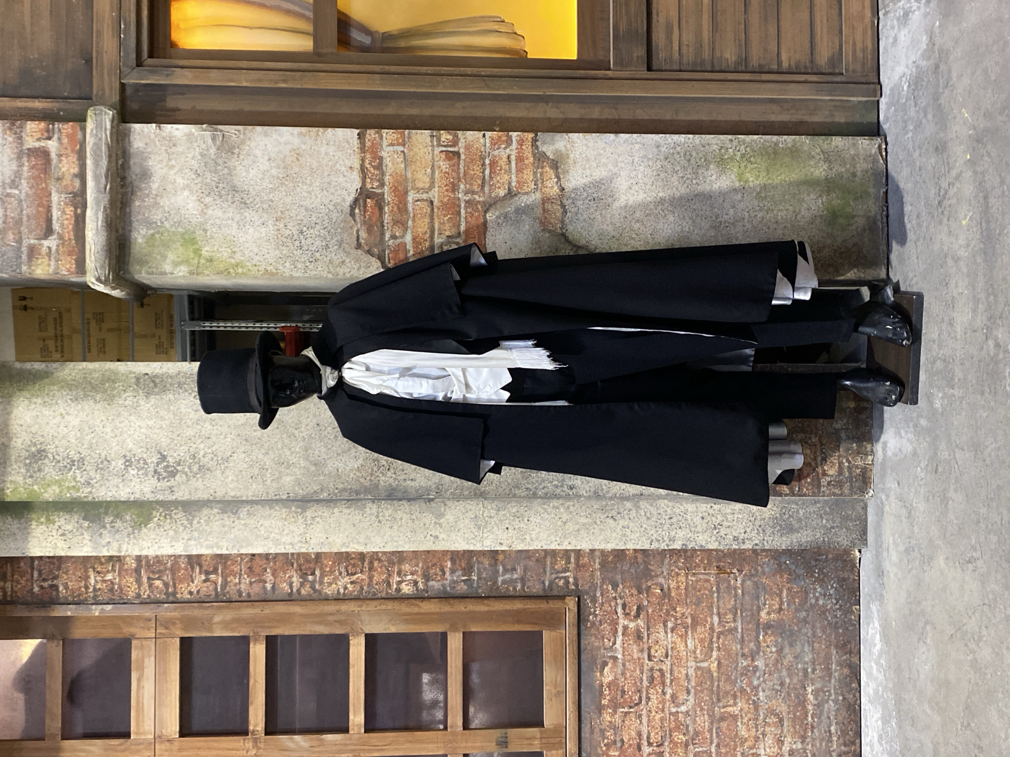 The image, rotated 90 degrees clockwise, displays a mannequin dressed in formal attire against a detailed, aged building facade. The mannequin stands prominently, wearing a black top hat, a black tailcoat over a white shirt, dark trousers, and black shoes. Its head appears to be a dark, featureless mask or painted black.
The background forms an elaborate set piece, simulating an old urban building. The facade is characterized by alternating sections of exposed red brick and weathered grey concrete or plaster walls, which exhibit distinct patches of green moss or mildew, suggesting significant age and exposure to the elements. Dark wooden window frames are visible at both the top and bottom of the frame. Through an upper window, a bright yellow light or object is visible, hinting at an interior light source or a theatrical effect. A downspout or pipe-like structure runs vertically on the left side of the building. In the upper-left background, behind the facade, cardboard boxes and a metal shelving unit are faintly visible, indicating a backstage or storage area beyond the display.
This is a static scene, depicting no activity. The historical and worn details of the facade, combined with the mannequin's period-appropriate attire, evoke a sense of a late 19th or early 20th-century urban environment. Given the location context of Buenos Aires, Argentina, this could be part of a museum exhibit, a theatrical set, or a themed display within the city. No discernible text is visible on the mannequin or the building elements.