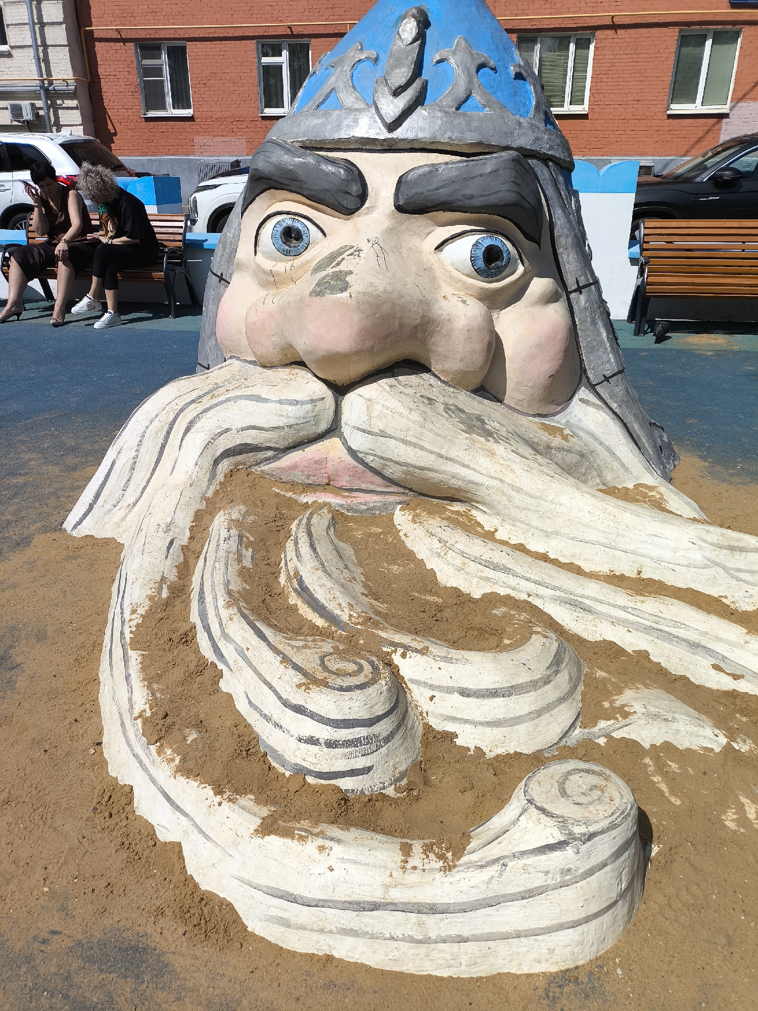 The image shows a large, painted sculpture of a bearded man's head wearing a blue and grey hat. The sculpture appears to be partially buried in sand. In the background, two women are sitting on a bench. There are parked cars and a brick building with windows. The location is in an urban environment. The city and country are unknown.