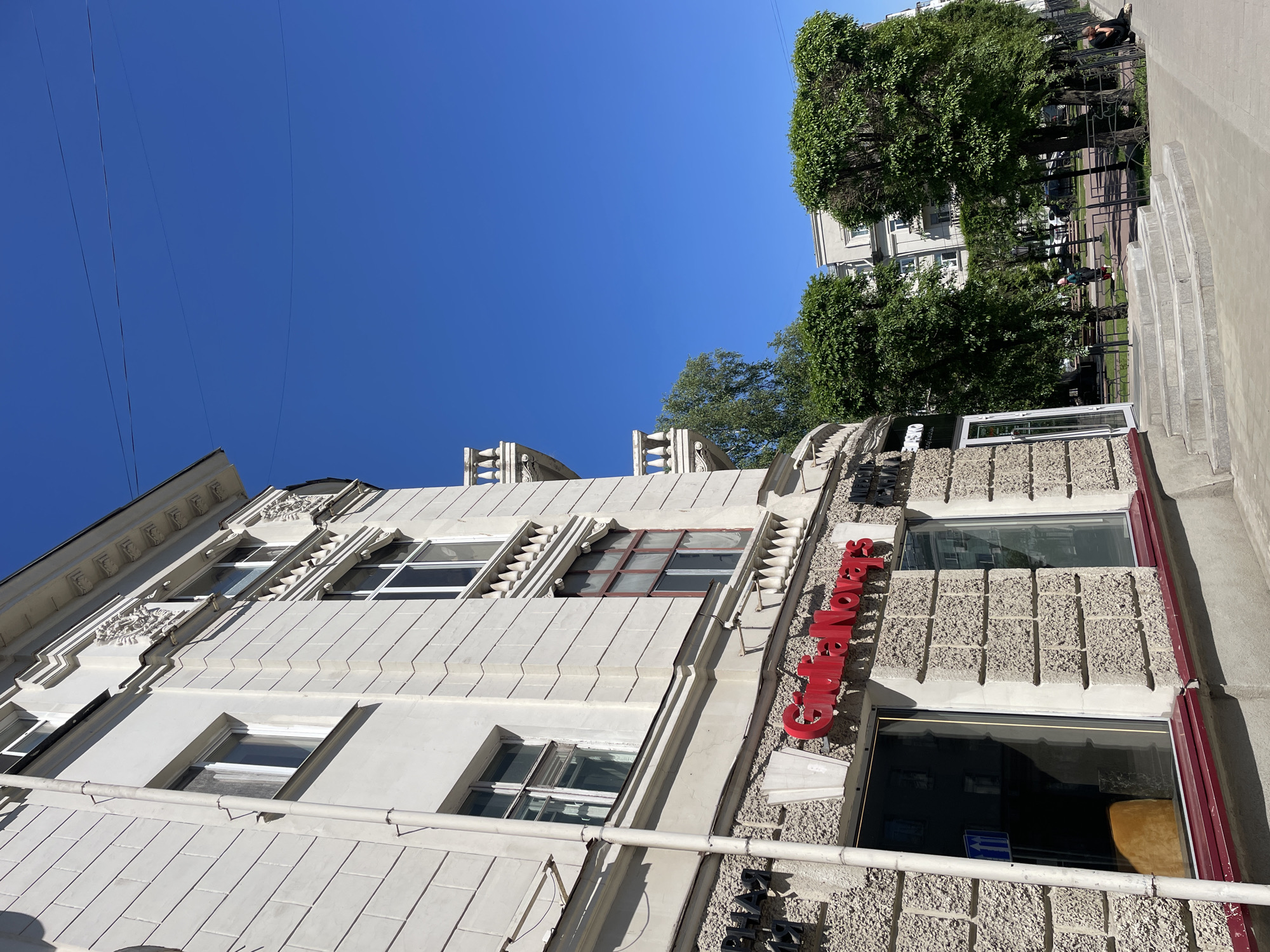 The image, originally rotated counter-clockwise, captures a sunlit urban street scene in Krasnoyarsk, Russia, featuring a building facade, a tree-lined street, and clear blue sky.
On the left, a multi-story building presents a white, ornate upper facade with intricate decorative architectural elements, including elaborate window frames, cornices, and friezes. Below this, the building's exterior transitions to a textured grey concrete block pattern. A prominent red sign with raised letters spelling "Gusta Novas" is affixed to this lower section. Below it, a dark sign with illegible text is visible, along with a blue sign on a windowpane.
To the right, a wide concrete sidewalk with ascending steps leads towards a row of dense, neatly trimmed green trees. These trees partially obscure a street with parked cars and other buildings in the background. Two individuals are visible on the sidewalk: one seated on a bench or railing, and another standing further down near a light pole.
The sky is a clear, vibrant blue, with several thin overhead wires crossing the upper left portion of the frame. The bright sunlight casts distinct shadows, indicating it is likely mid-day. The weather appears sunny and clear.