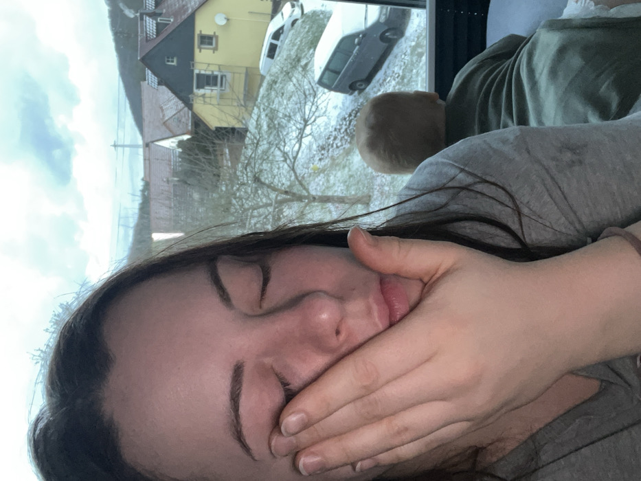 A woman is reclined, facing the camera. Her eyes are closed and a hand is covering her mouth. Behind her is a window showing a snowy scene with houses, trees, and vehicles covered in snow. A light-haired baby's head is visible between the woman and the window. The woman wears a grey shirt, and a green shirt is also visible.
