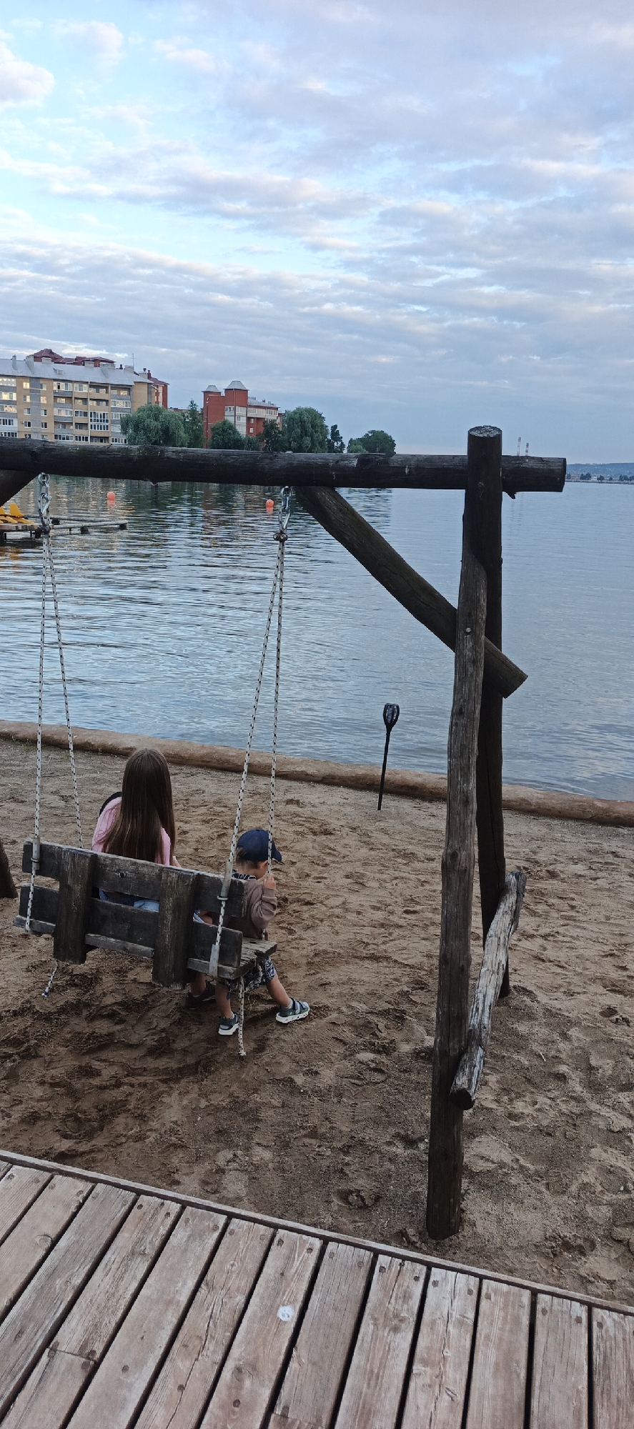 The image depicts two children on a wooden swing by the shore of a body of water, possibly a lake or river. In the background, a city skyline is visible, featuring apartment buildings and trees. A wooden pier is in the foreground. The sky is cloudy, and the overall lighting suggests it is either early morning or late evening. The location is identified as Votkinsk, Russia. The swing set is constructed from rough-hewn logs and has rope supports. The children appear to be a girl with long brown hair wearing a pink shirt and denim shorts, and a younger boy wearing a blue baseball cap, a brown shirt, and patterned shorts. They are seated on the swing, facing away from the camera. The ground is sandy, with a wooden boardwalk to the left.